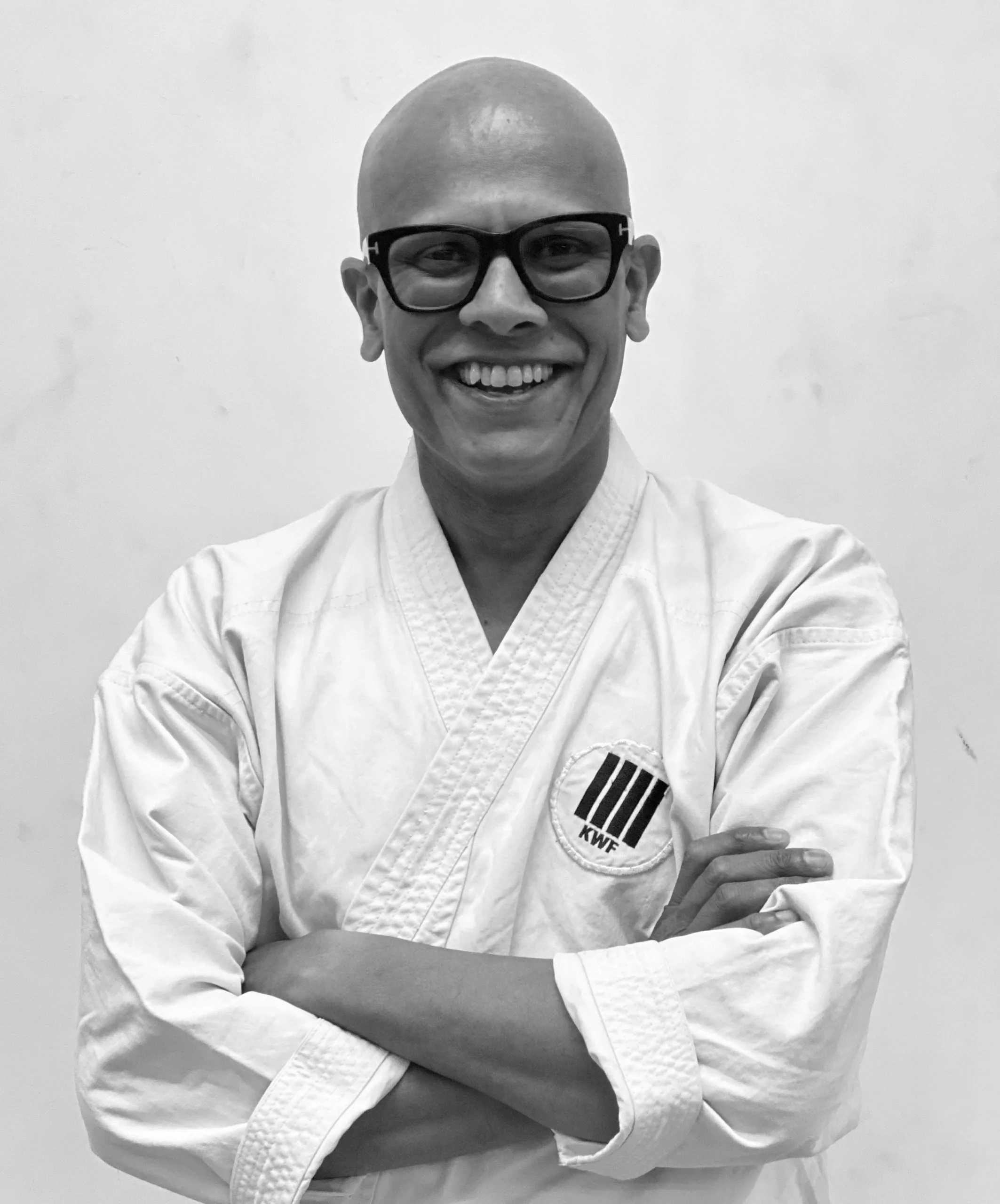 A person wearing glasses and a martial arts uniform with a KWF logo, smiling and crossing arms, against a plain background.