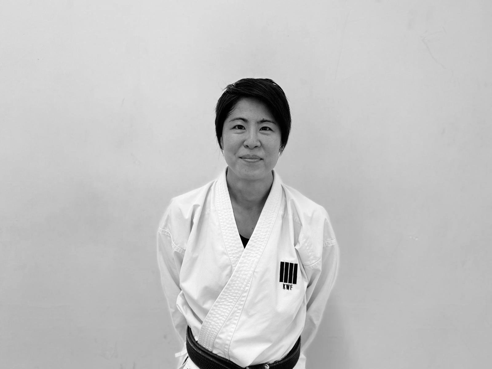 A woman with short dark hair wearing a white martial arts gi with a black belt standing in front of a plain white wall.