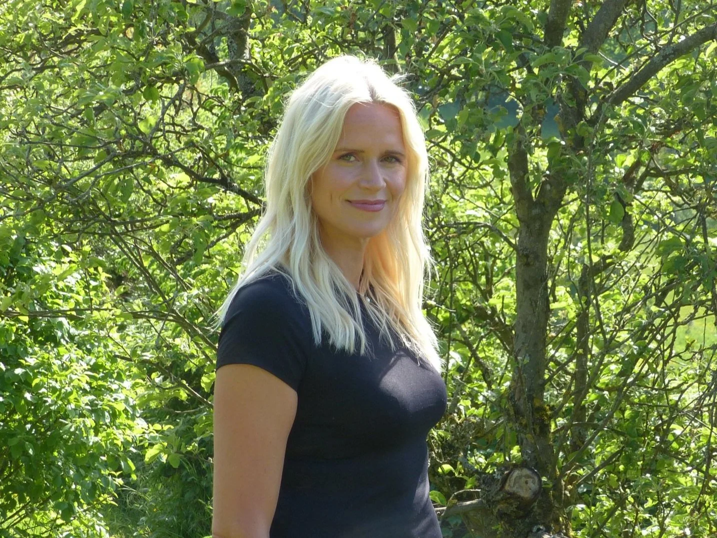 Eine blonde Frau steht in einem grünen Wald oder Garten.