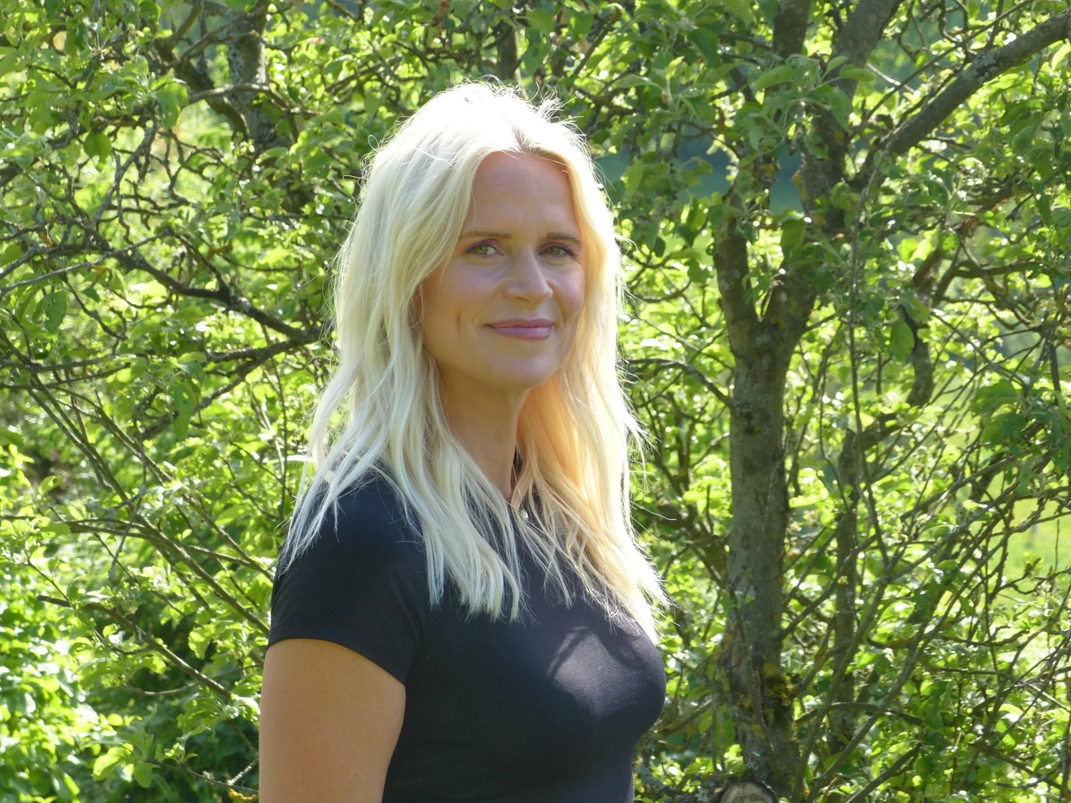 Blonde Frau in schwarzem T-Shirt steht vor grünem, belaubten Baum im Freien.