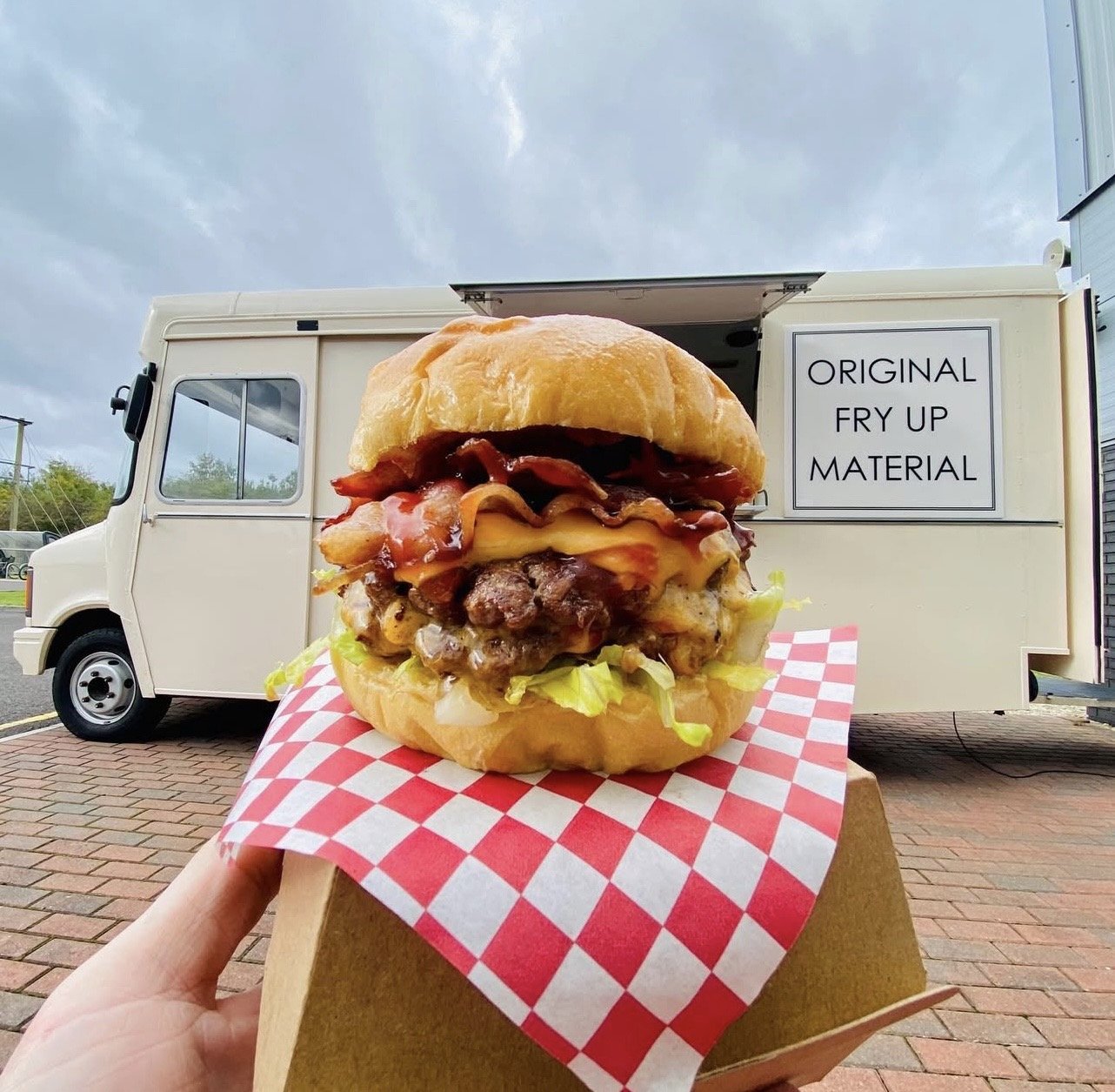 Close-up of a large cheeseburger with bacon, lettuce, tomato, cheese, and a bun, held on checkered paper in front of a food truck with a sign that reads "Original Fry Up Material". The truck is parked on a brick surface with a cloudy sky in the background.