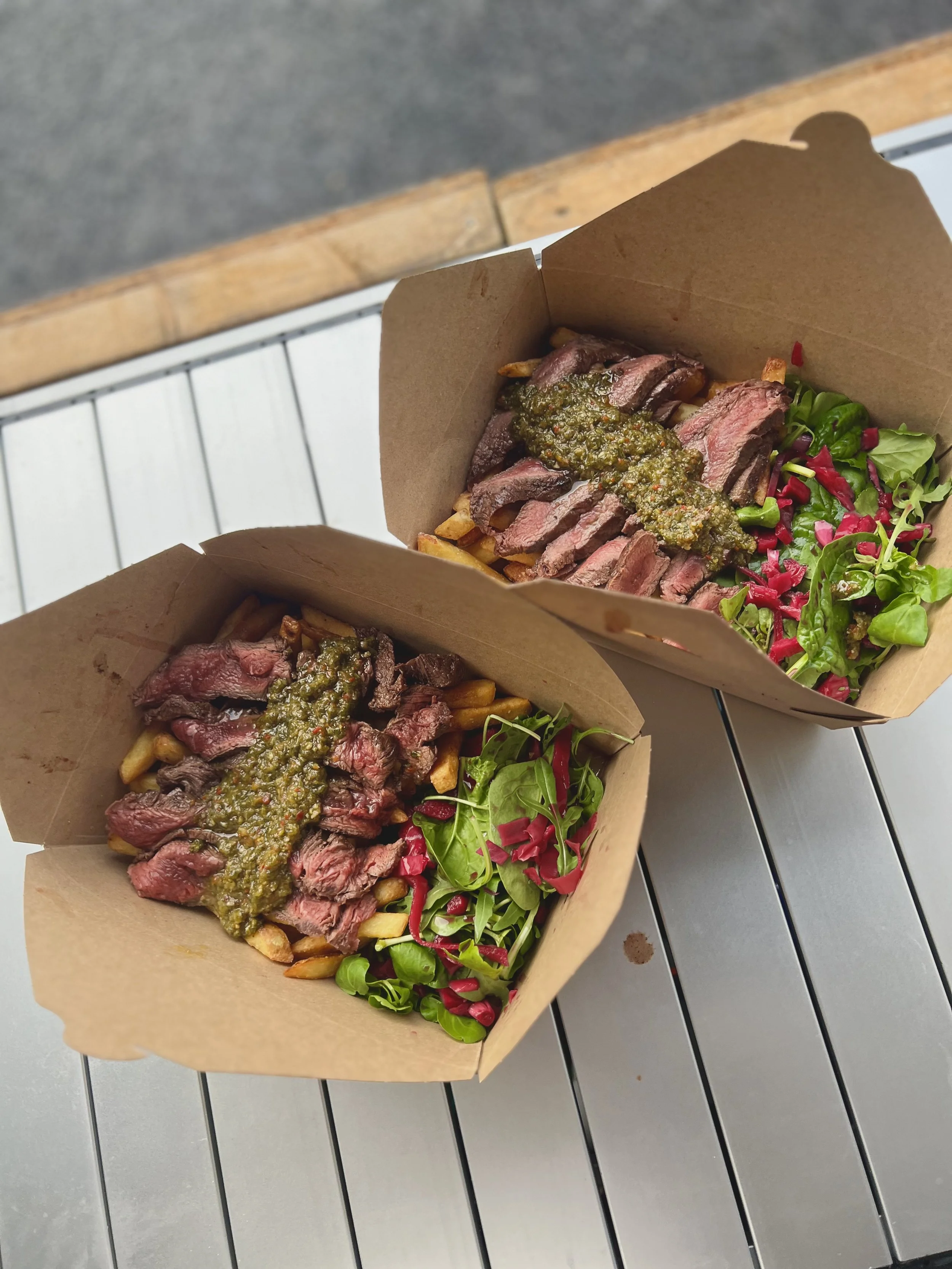 Two cardboard trays of French fries topped with slices of beef steak and sauce, garnished with greens and pink pickled vegetables, alongside a bowl of pink pickled vegetables.