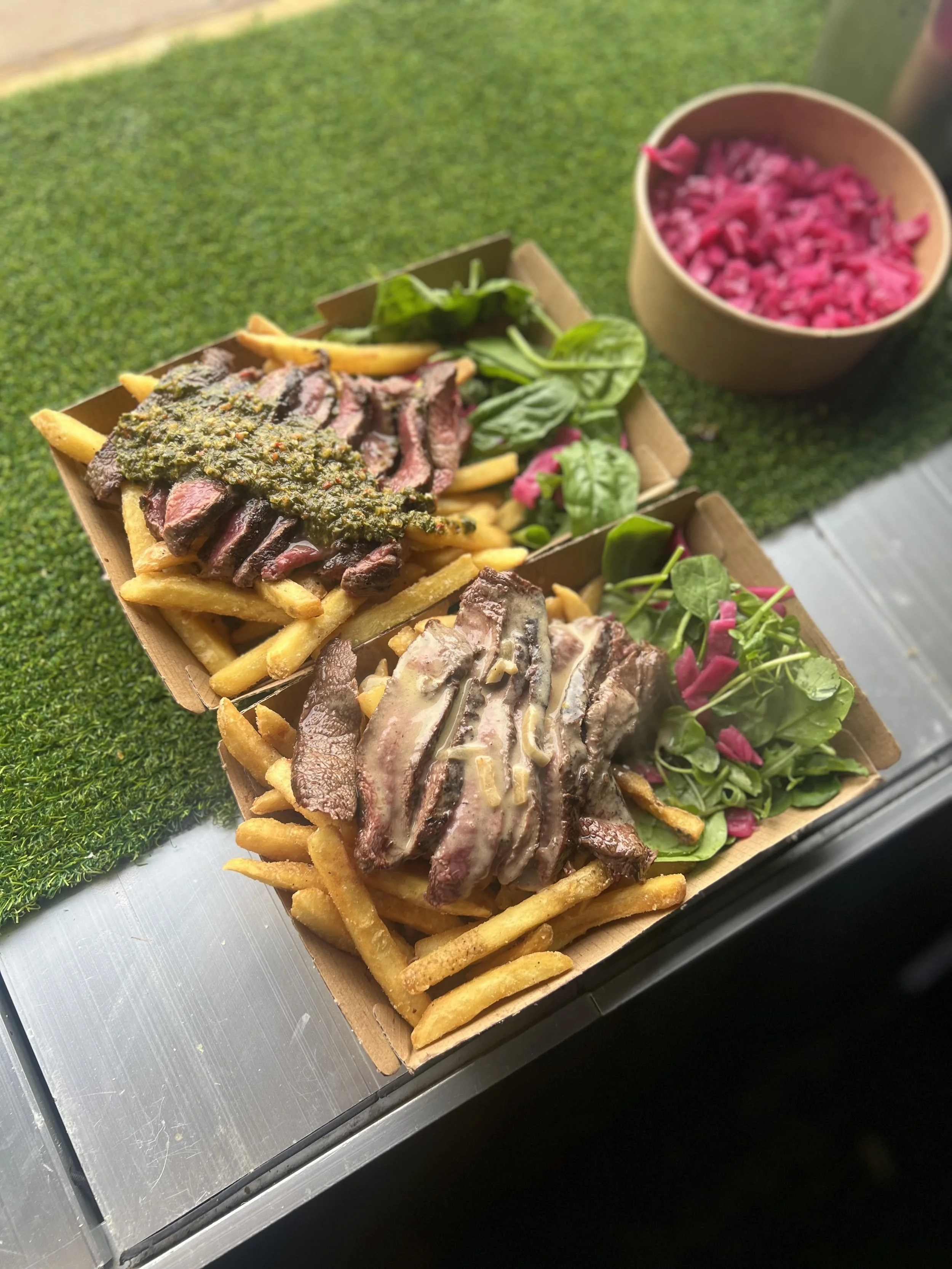 Two cardboard trays of French fries topped with slices of beef steak and sauce, garnished with greens and pink pickled vegetables, alongside a bowl of pink pickled vegetables.