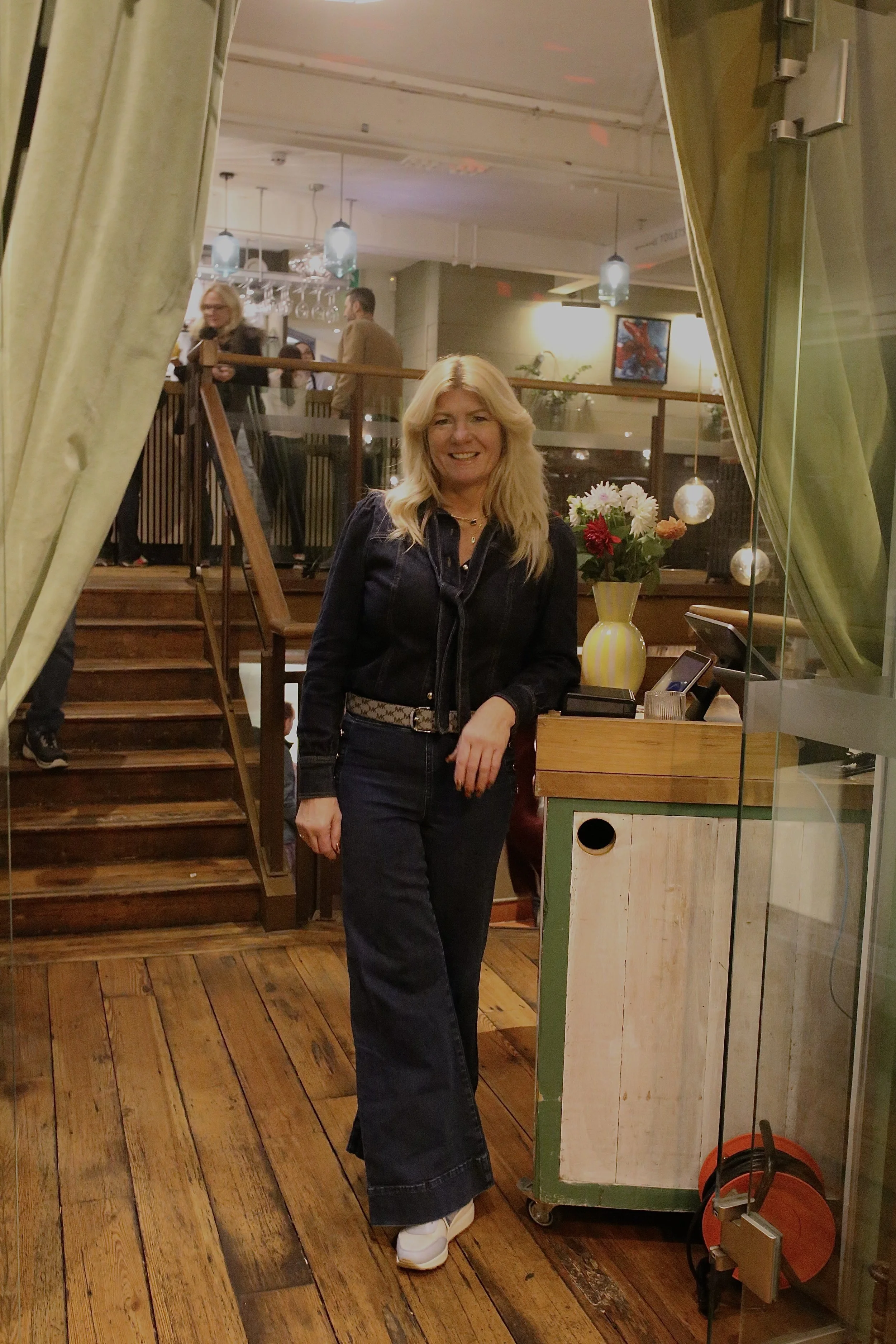 A woman with blonde hair smiling and posing indoors near a flower vase, with a staircase and people in the background.