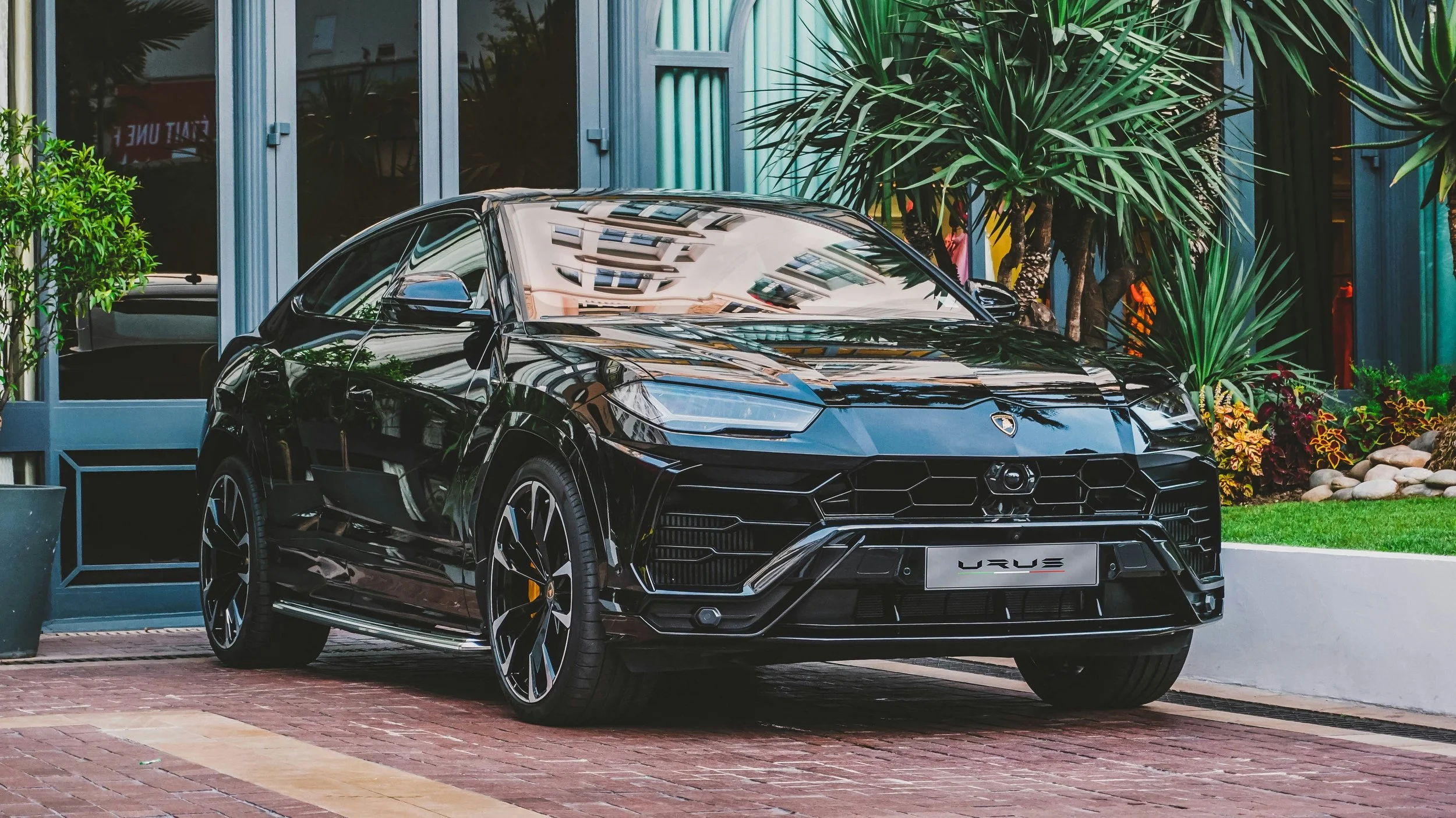 A black Lamborghini Urus parked outside a modern building with large glass windows, surrounded by green plants and a brick-paved sidewalk.