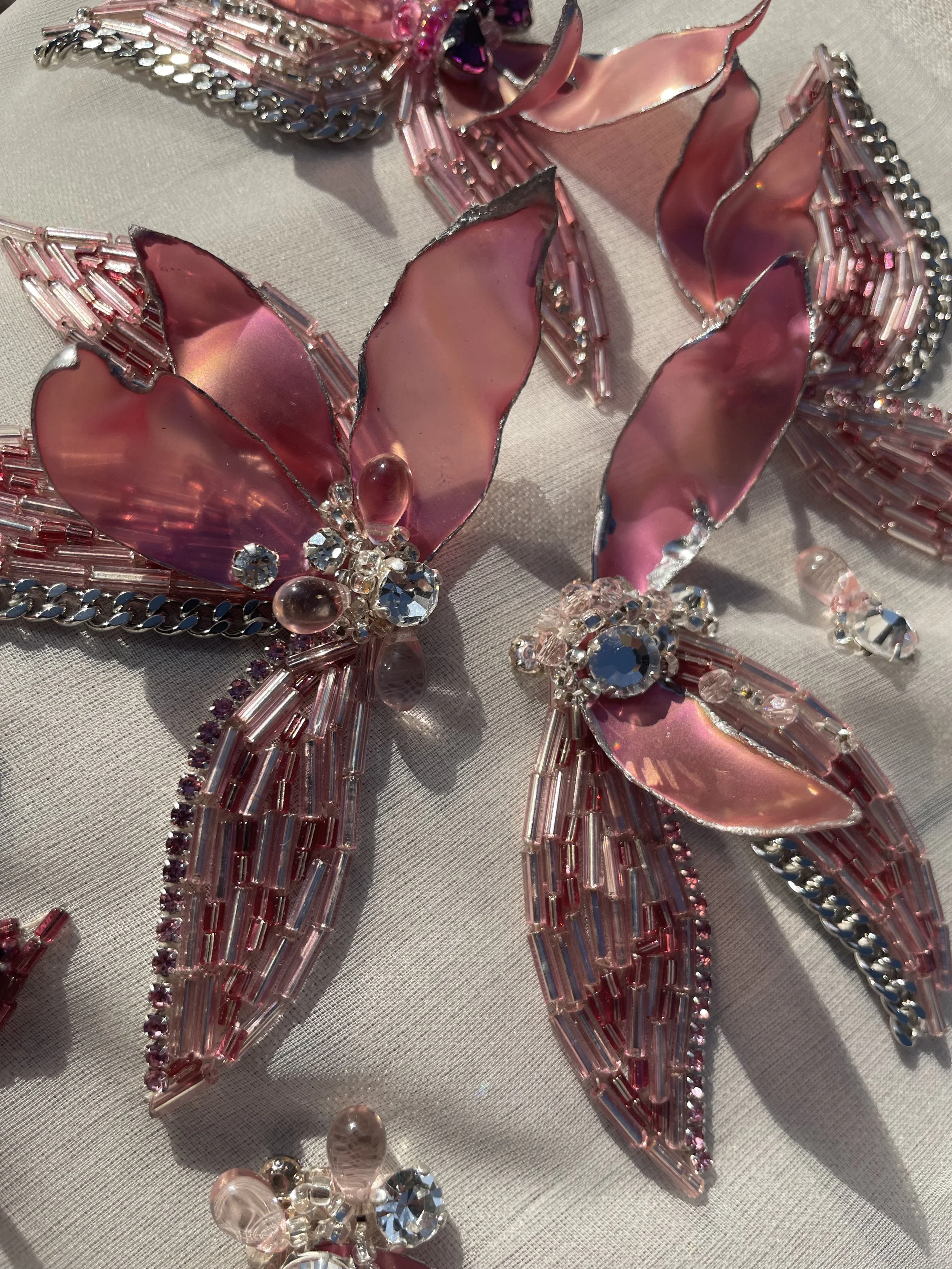 A decorative pink and silver butterfly brooch embellished with pink and clear rhinestones and beads, resting on a light fabric background.