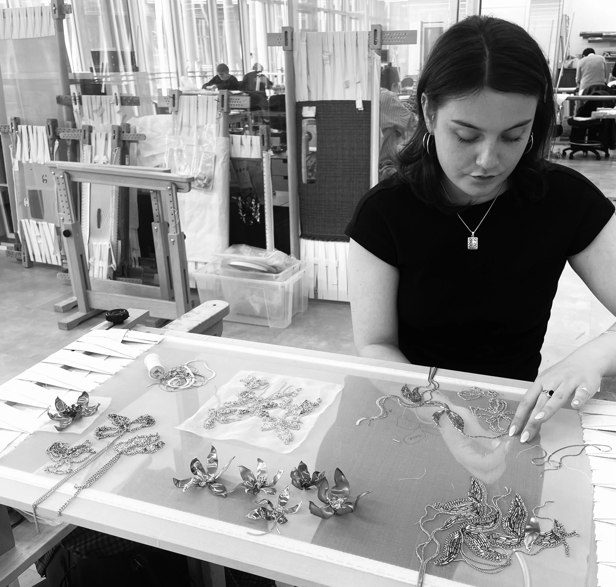 A woman with dark hair, wearing earrings and a necklace, is examining jewelry displayed on a table. The jewelry includes necklaces, bracelets, and earrings, and there are more jewelry items on the table. In the background, there are other people working and organizing items in what appears to be a workshop or studio with shelves and partitions.