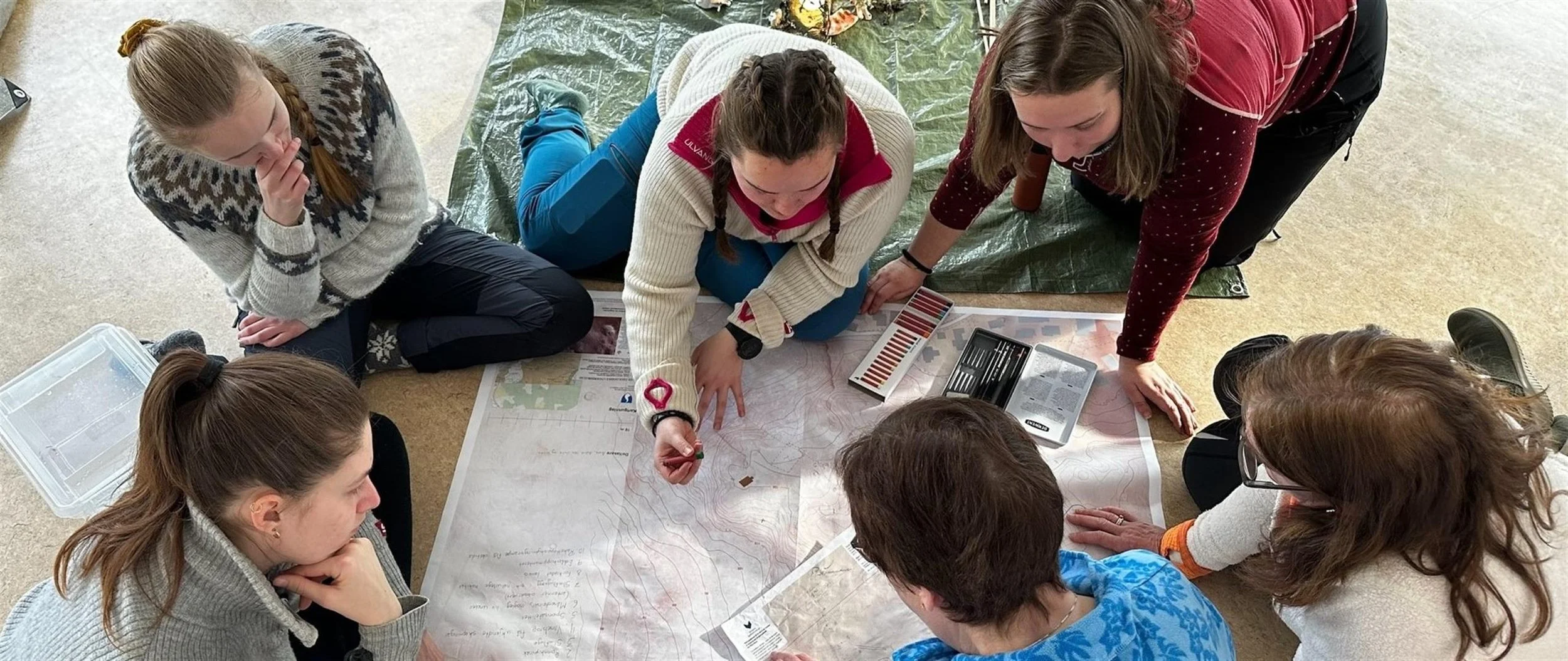 Grupp av personer som studerer et kart på gulvet, omgitt av utstyr som fargeprøver, linjaler og notatblokker, på innsiden.