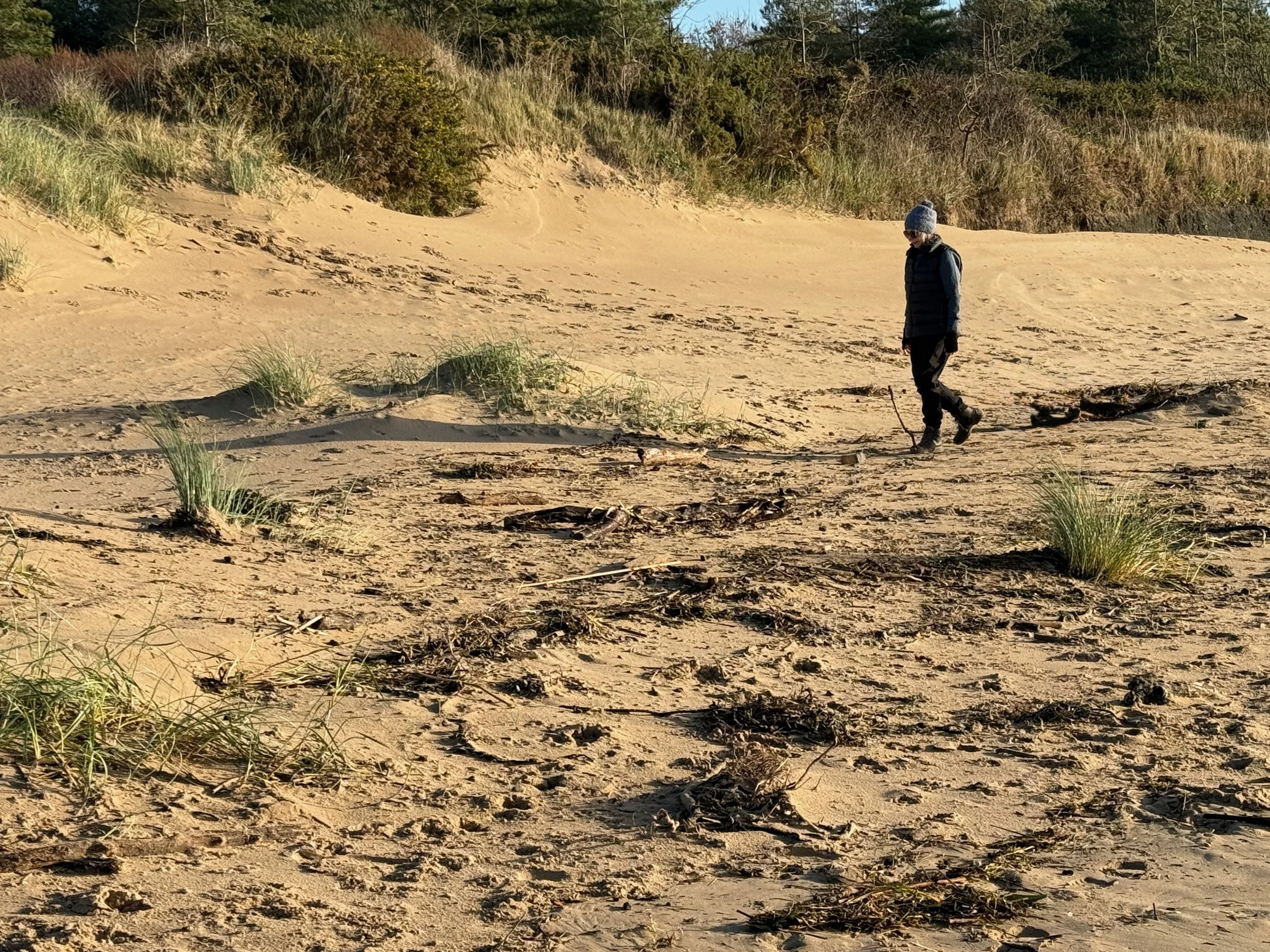 Roz Moreton walking along a the Post Industrial Landscape of the Carmarthen Bay Power Station with dunes and vegetation in the background, wearing a grey beanie, dark jacket, and work trousers.