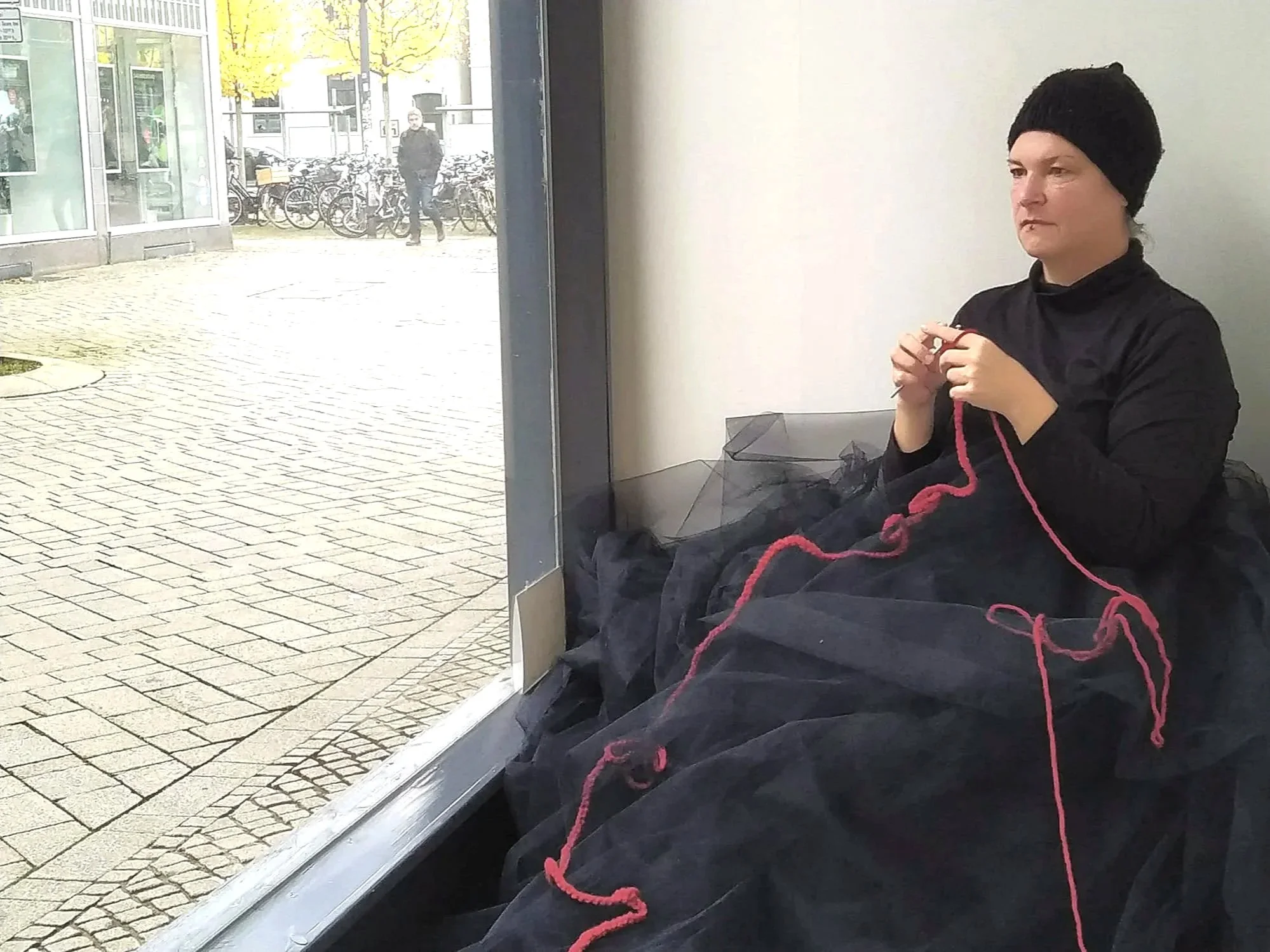Künstlerin sitzt häkelnd in einem Schaufenster, hält ein rotes Garn in den Händen. Im Hintergrund große Fenster mit Blick auf eine Fußgängerzone.