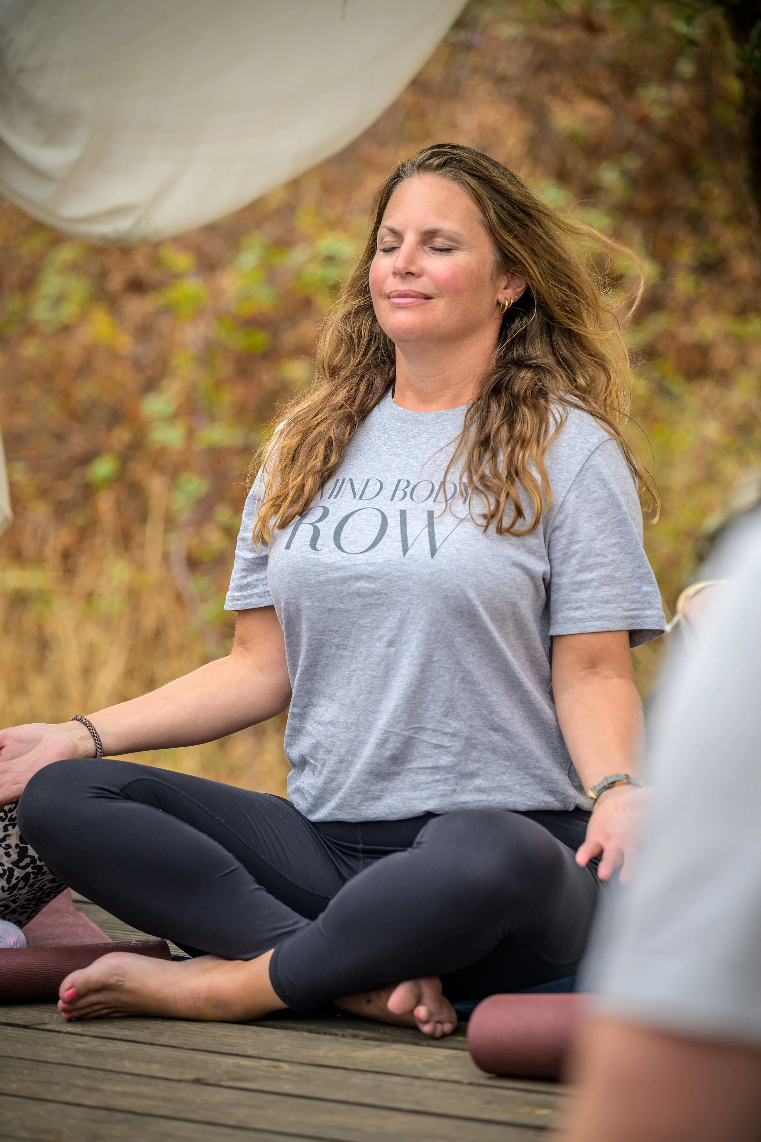 A woman practicing yoga outdoors on a wooden deck, sitting cross-legged with eyes closed, wearing a gray t-shirt and black leggings, surrounded by autumn foliage.