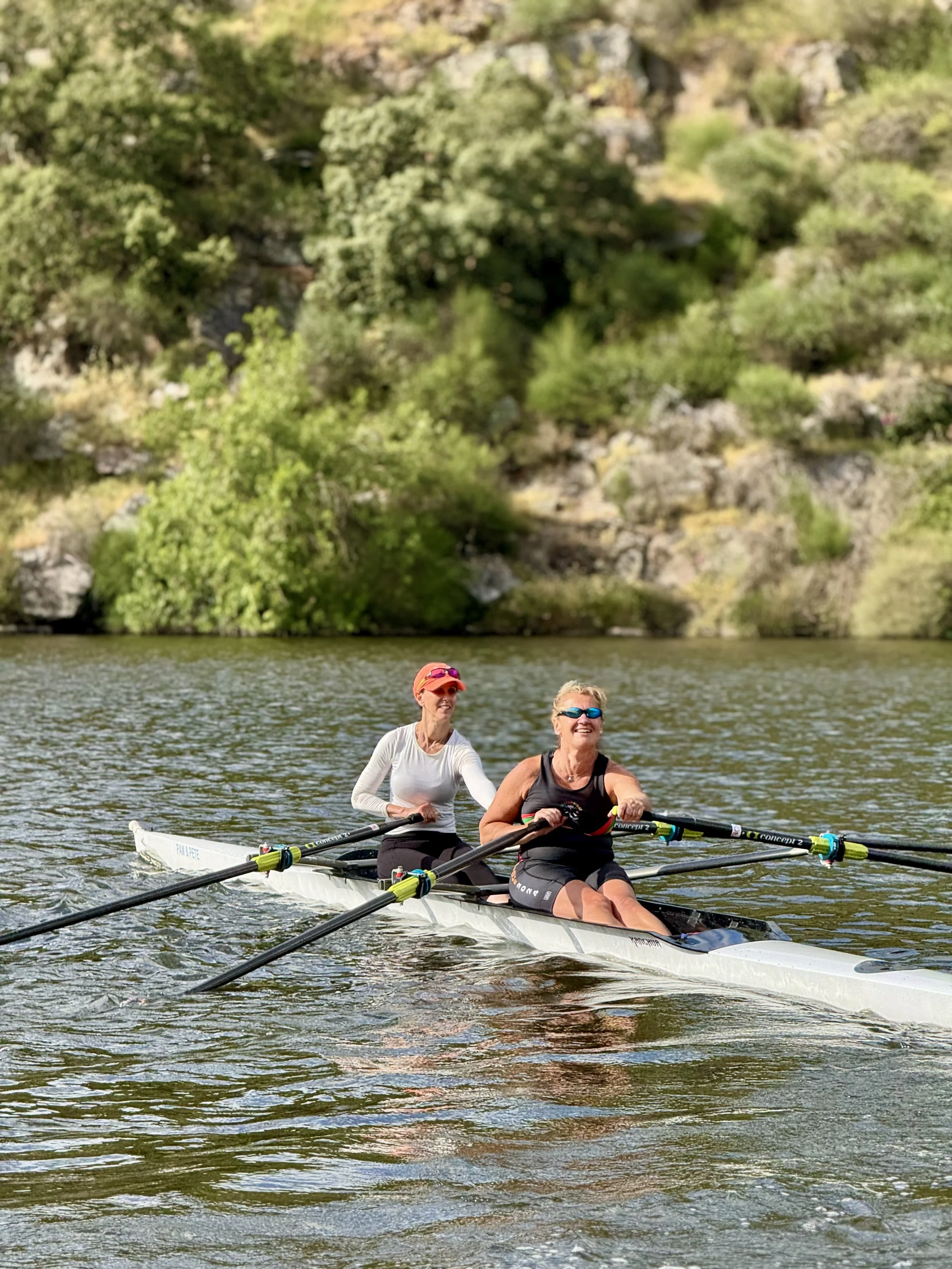 Two women rowing a boat on a lake with trees and rocky hills in the background.