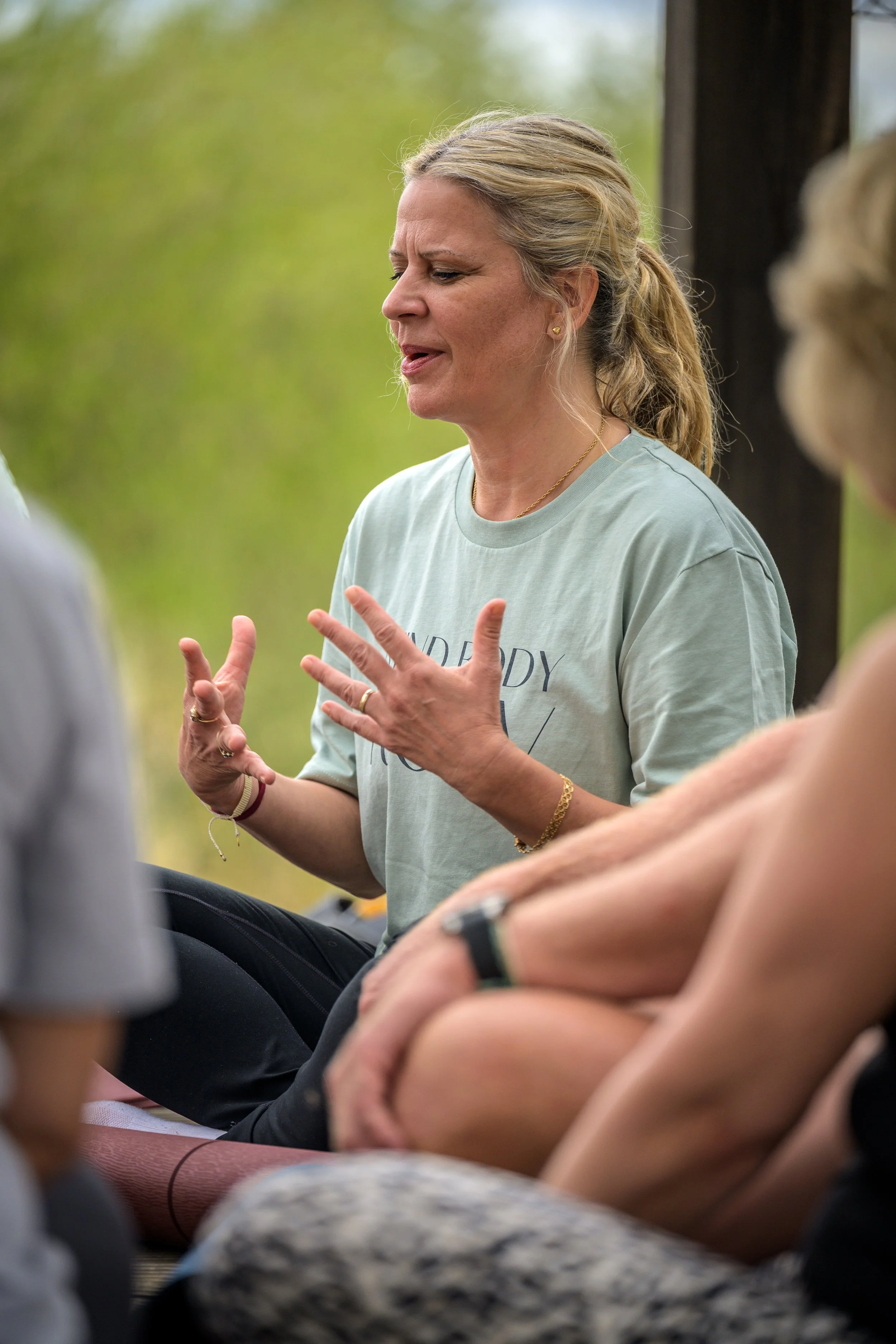 A woman is speaking to a group of people outdoors, using expressive hand gestures. The background is blurred greenery.