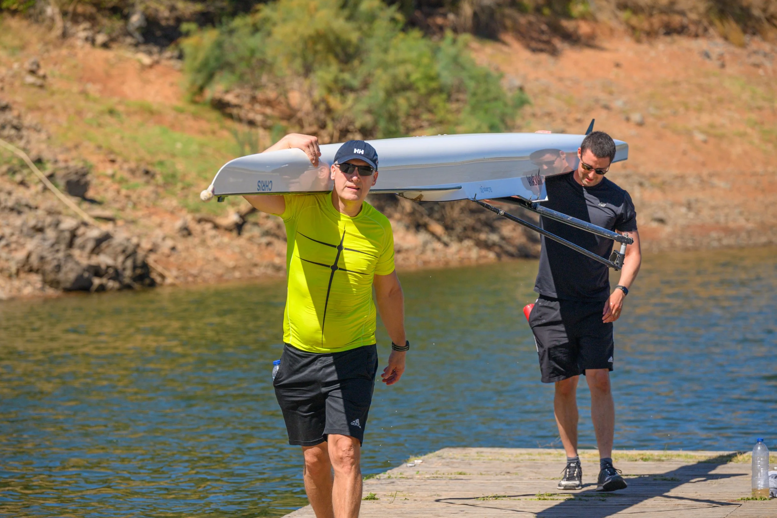 Two men in athletic clothing and sunglasses carrying a kayak on their shoulders along a wooden dock by a lake, with rocky shoreline and trees in the background.
