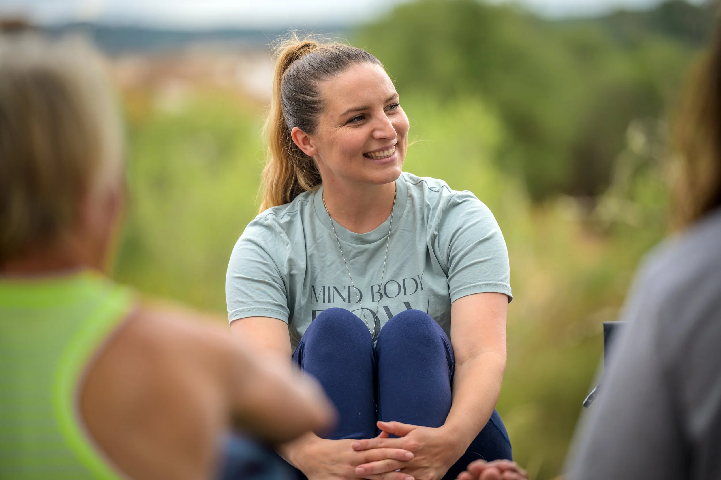 A woman with a ponytail smiling and talking to a group outdoors, with trees and greenery in the background.