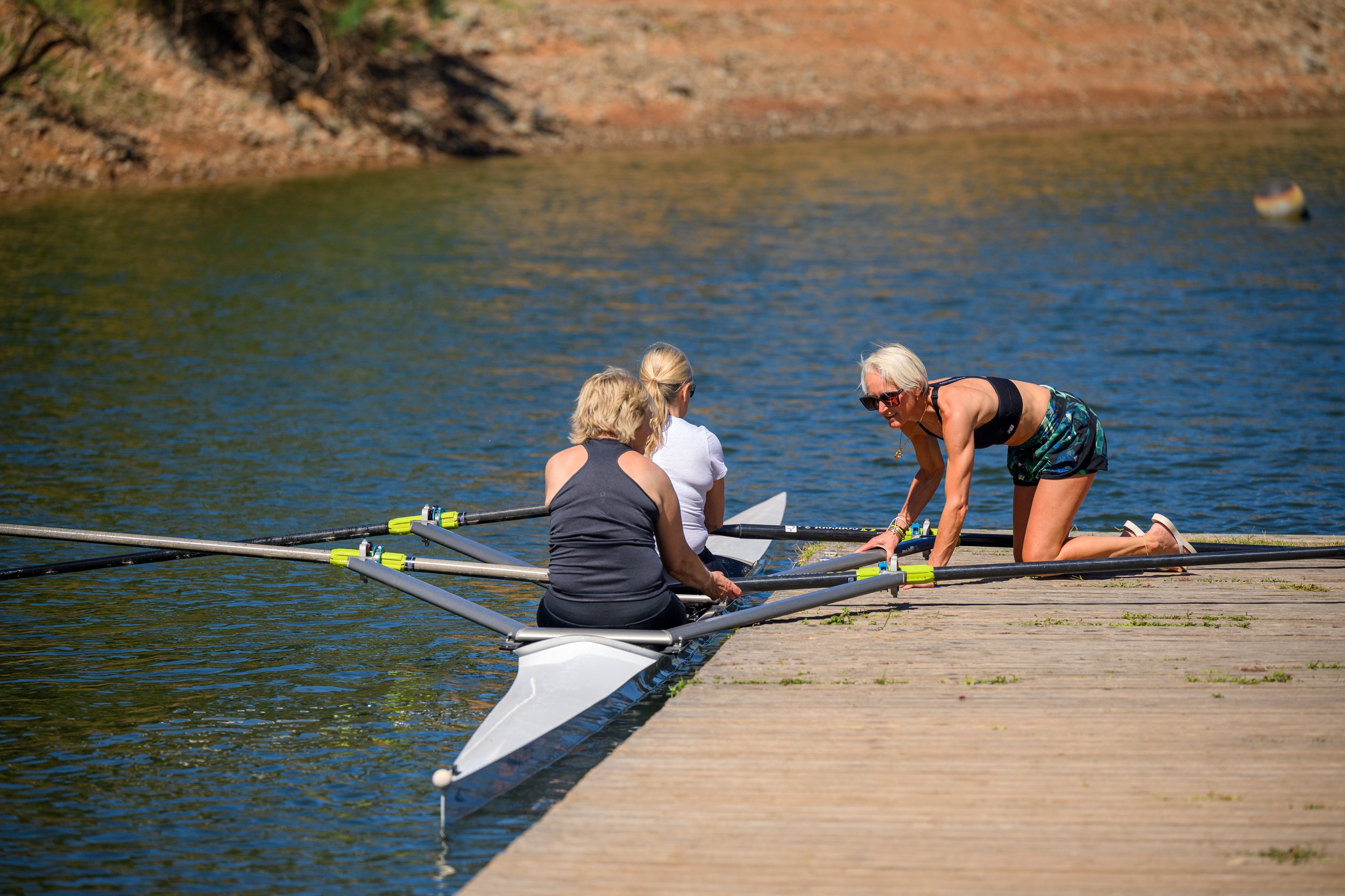 Three women preparing a rowing boat at the edge of a lake or river, with one woman kneeling on the dock and others sitting inside the boat.