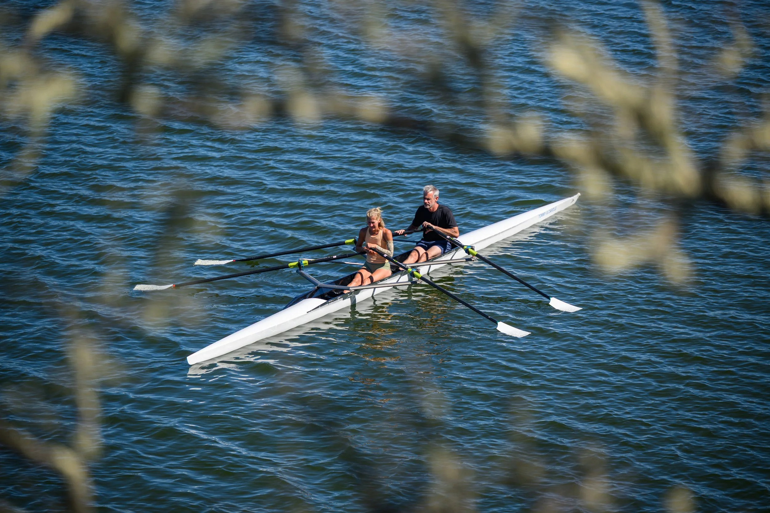 A man and a woman row a sculling boat on a river, viewed through branches of a tree.