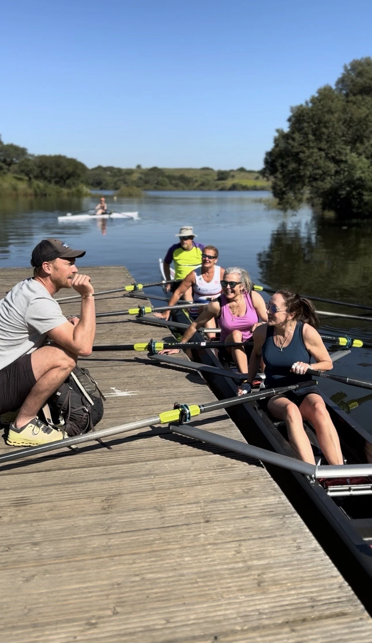 A group of people preparing to row a boat, sitting on a dock by a calm river under clear blue skies, with a person kayaking in the background.