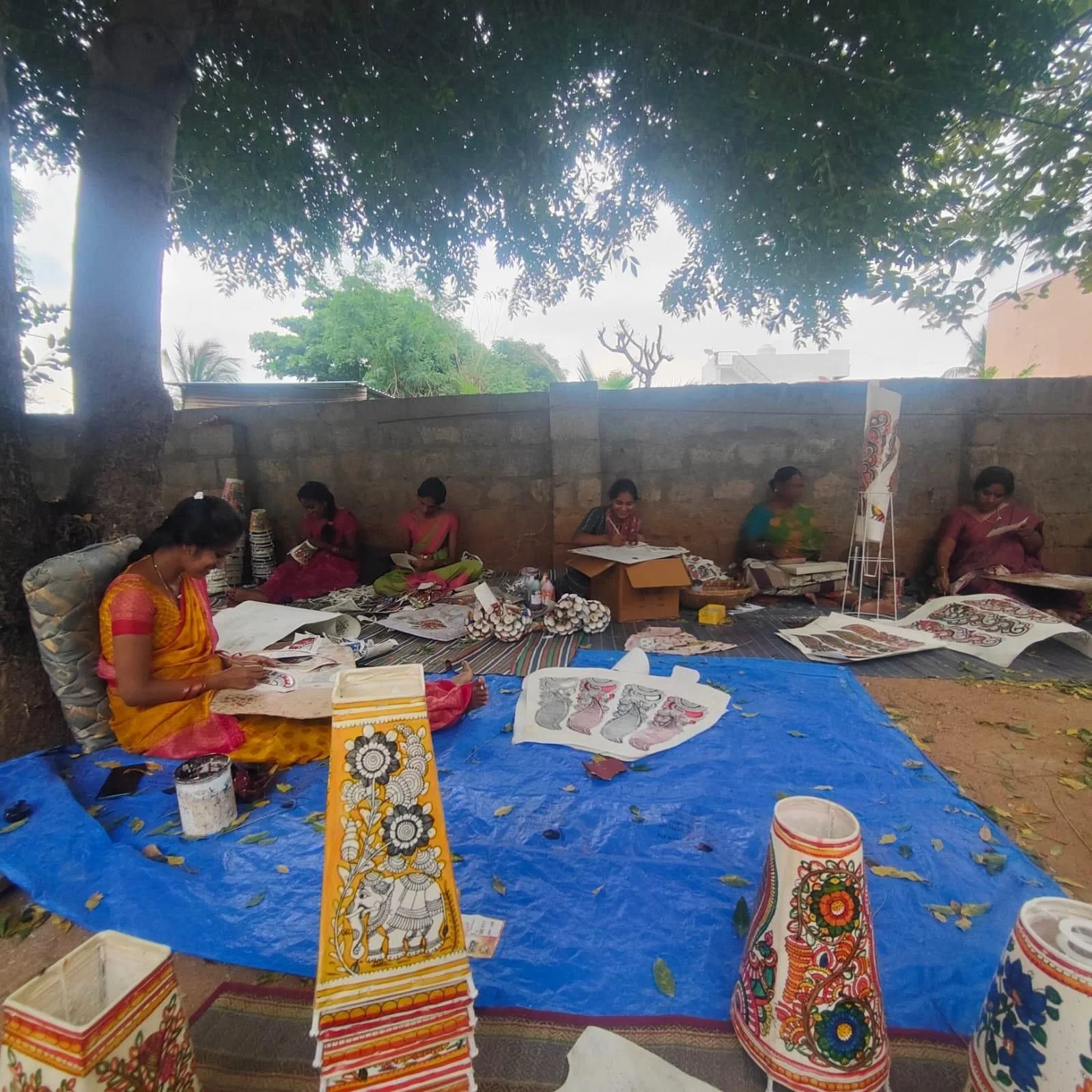 Six women sitting under a tree against a brick wall, engaged in traditional art or craft work, with colorful painted vases and sketches on display.
