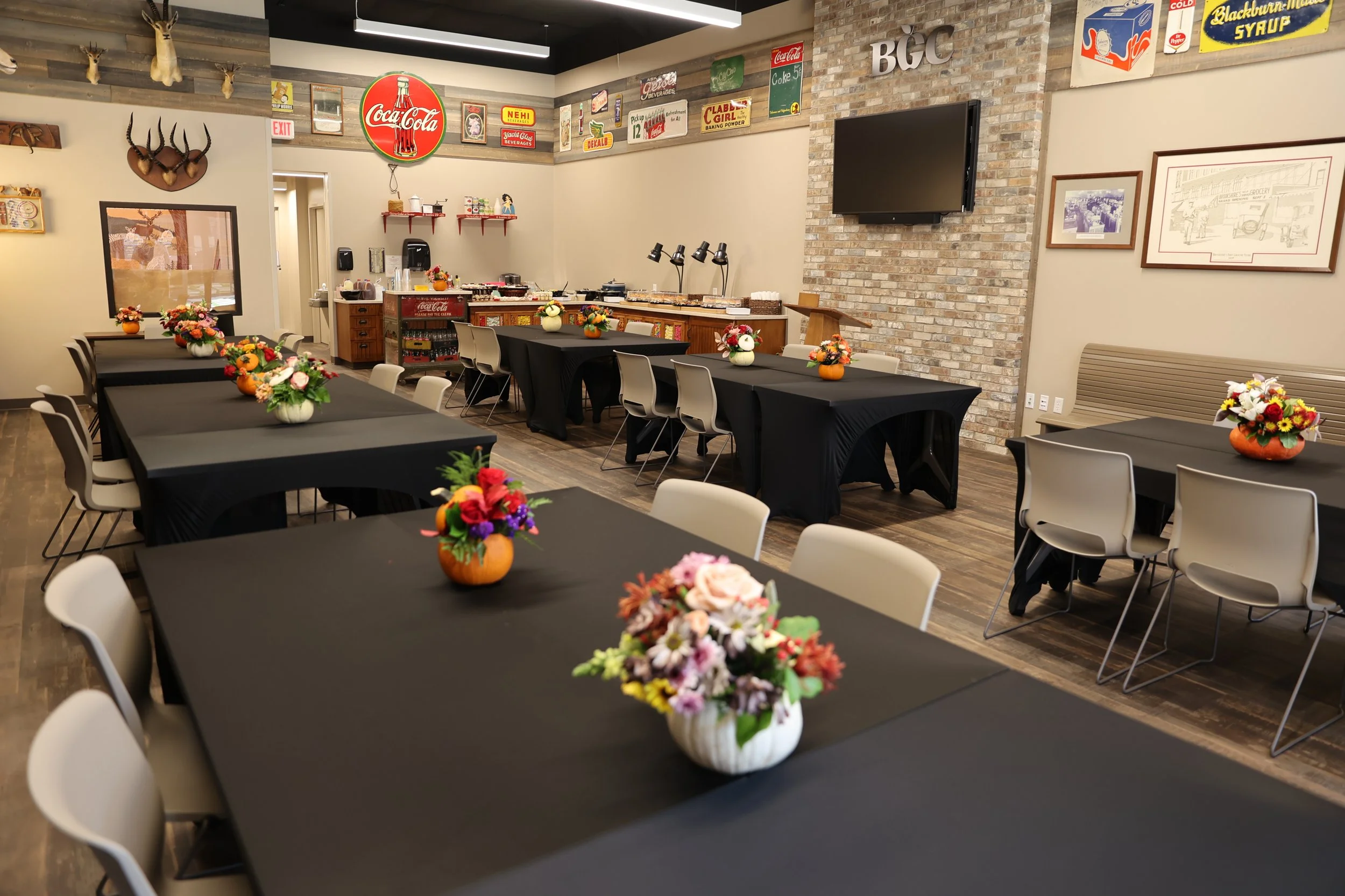 A banquet hall set up for an event with multiple tables covered in black tablecloths, each adorned with colorful flower arrangements in small pumpkins and vases. The room features a brick wall with a large TV, vintage signs, and décor, along with a b