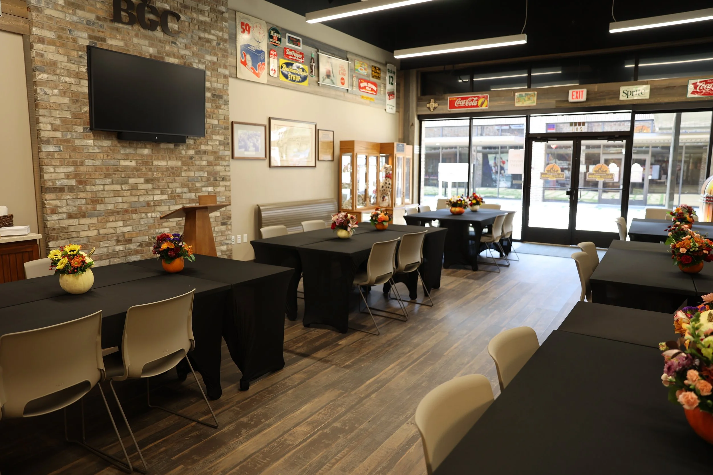 Empty indoor dining area with black tables, beige chairs, and flower arrangements, facing a glass storefront entrance with signs and decorations, and a brick wall with a TV mounted on it.
