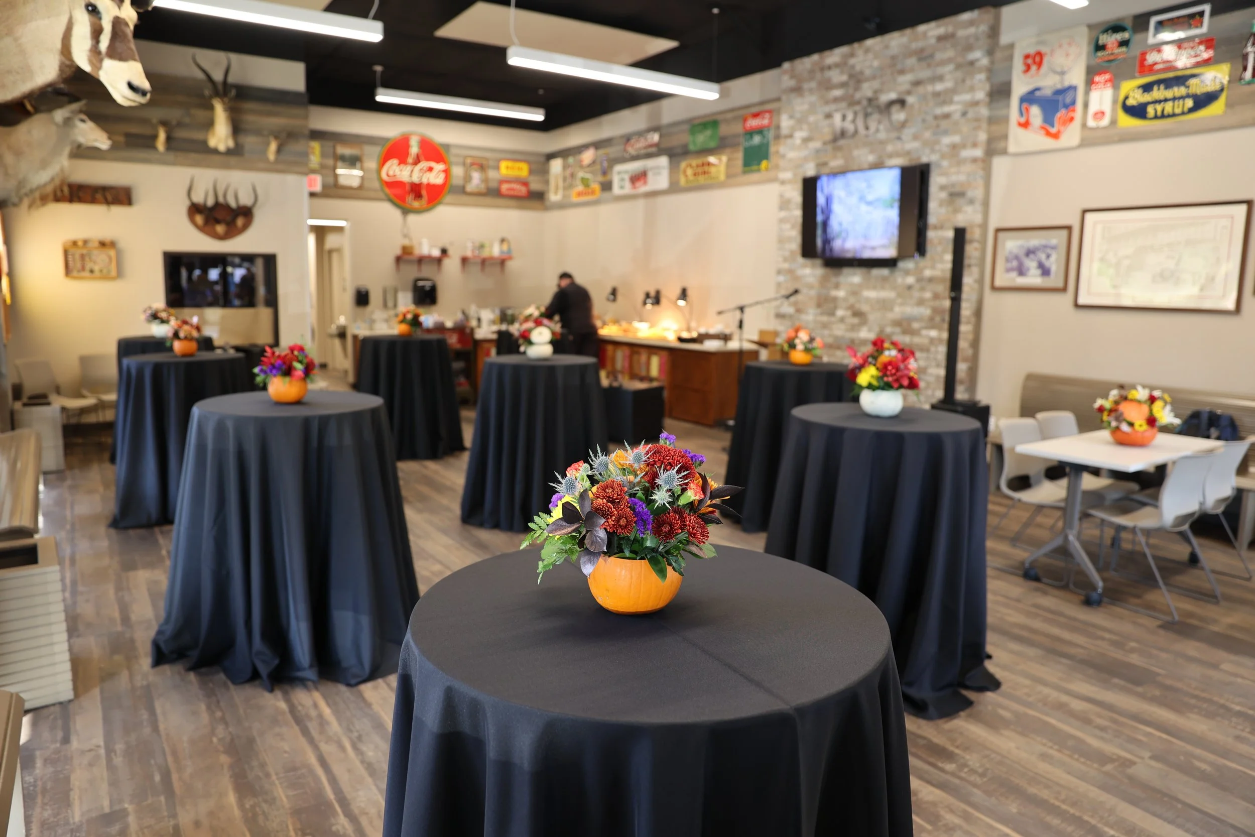 Room decorated with black tablecloth-covered tables, each with colorful flower arrangements, likely set up for an event or gathering, with a bar area and vintage signage decor on the walls.