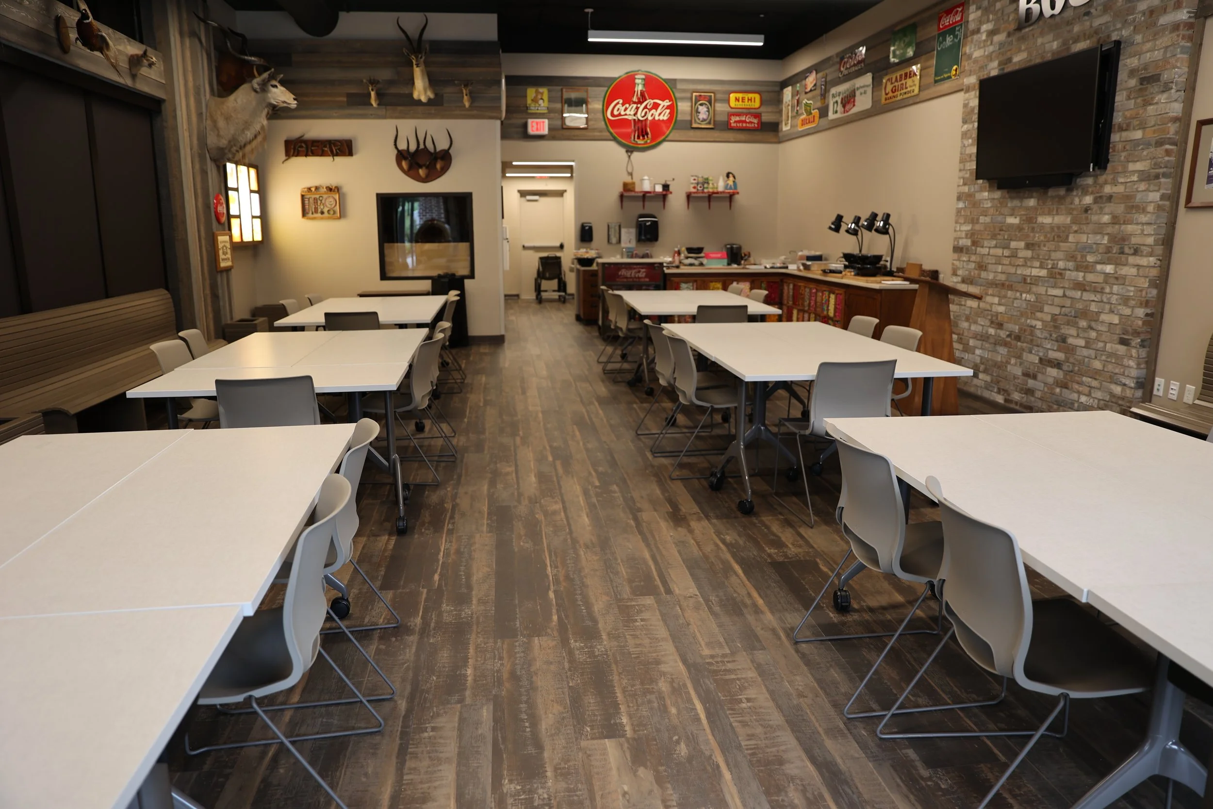 A modern dining area with white tables and gray chairs, featuring rustic wood flooring, a brick accent wall, with vintage signs and animal heads mounted on the walls, and a Coca-Cola sign on the back wall.