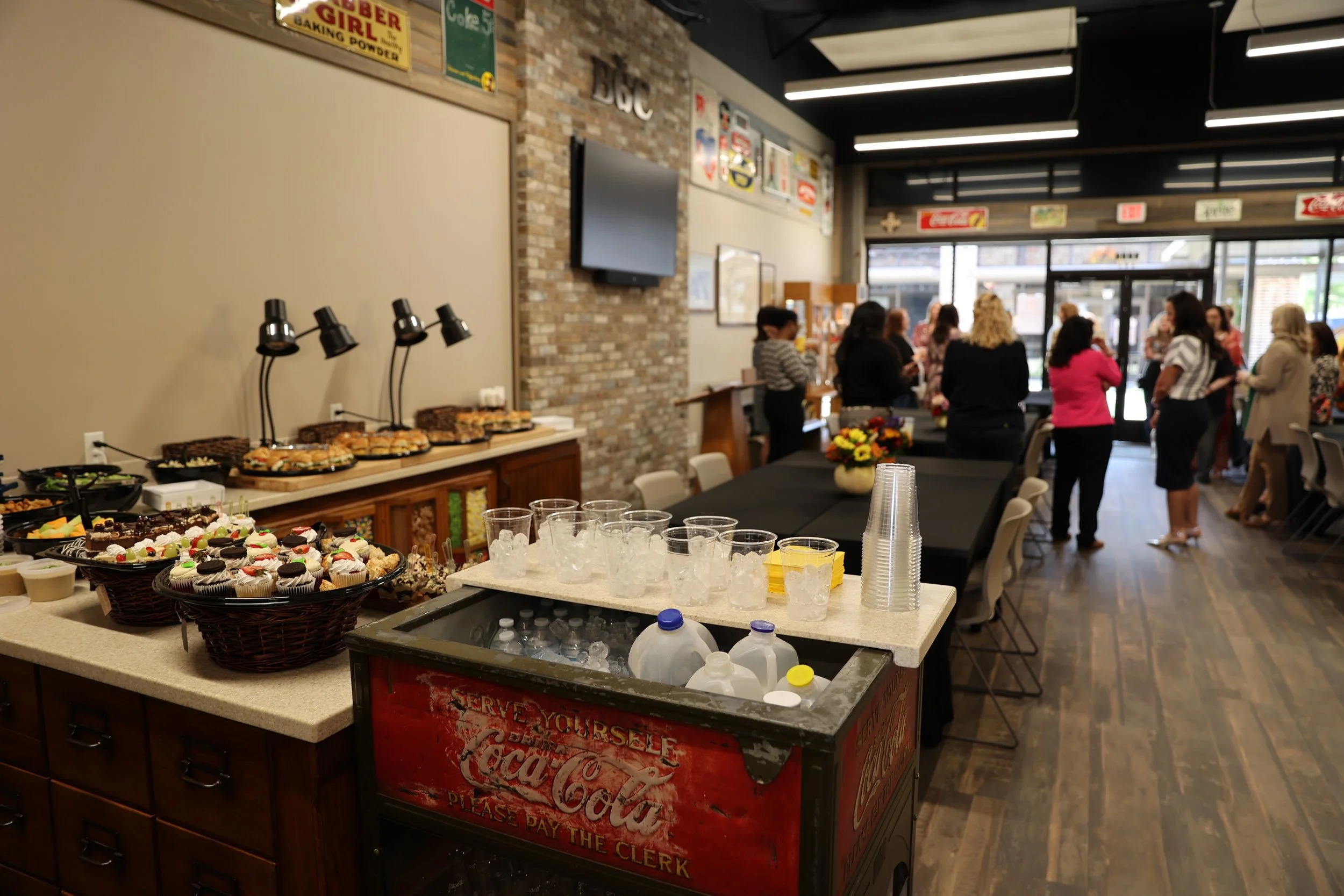Food and drinks setup at an indoor gathering with cupcakes, cookies, and beverages in cups and bottles, and people mingling near the entrance of a decorated room.
