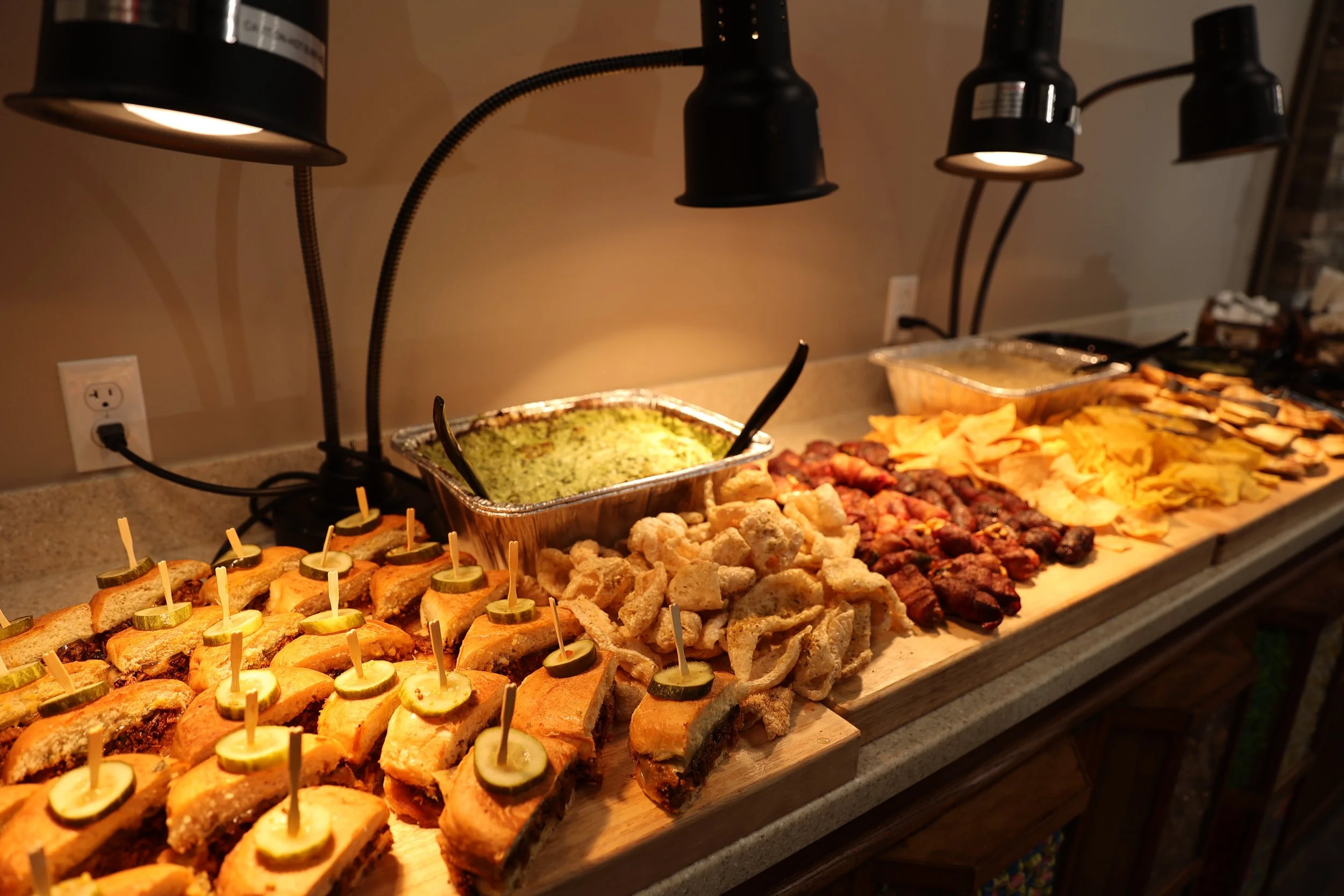 A buffet table with mini sandwiches topped with pickles, a tray of guacamole, fried pork rinds, skewered meat, potato chips, and grilled chicken in the background, illuminated by black adjustable lamps.