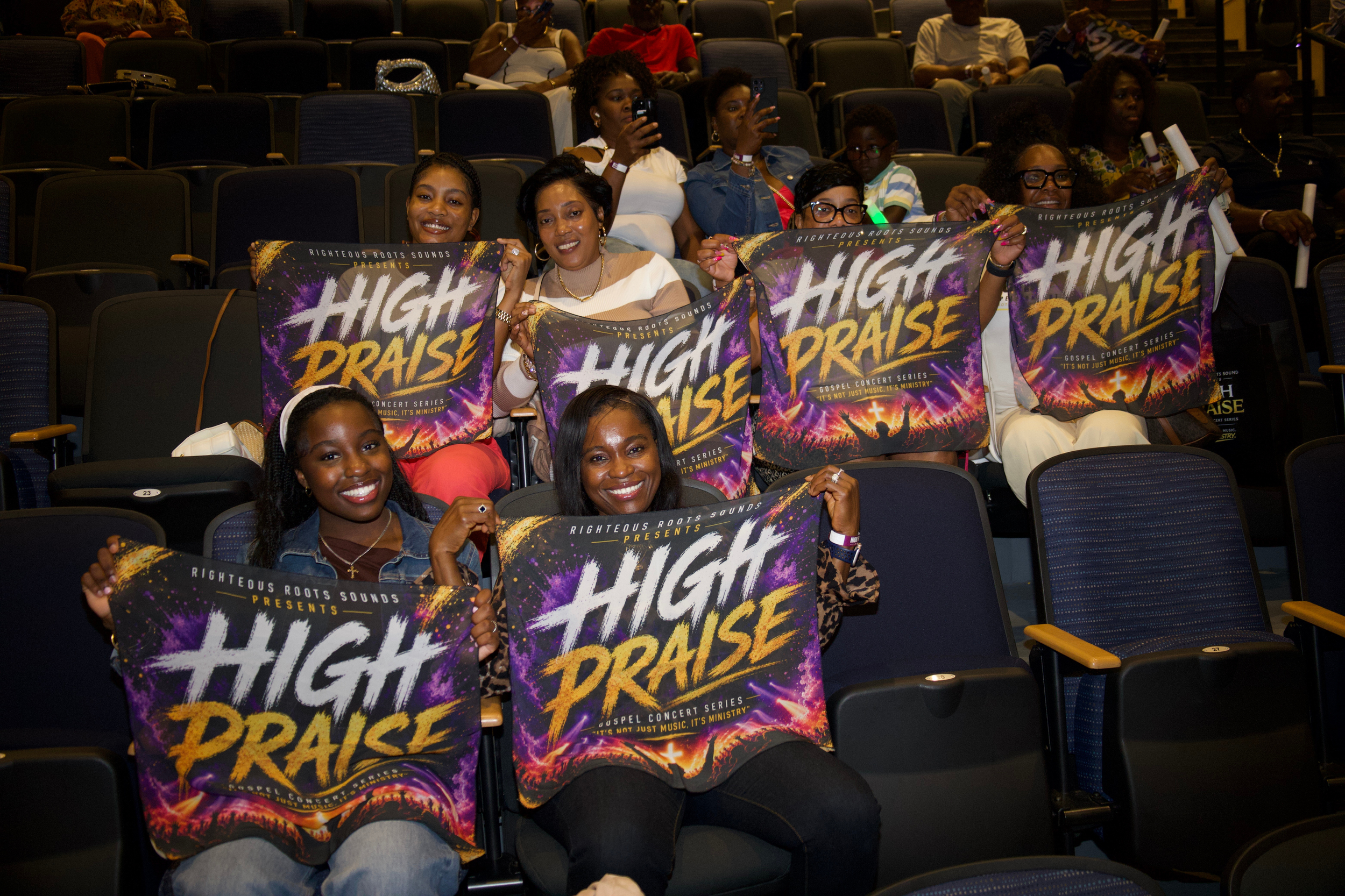 A group of women and girls sitting in an auditorium, holding up colorful signs that say 'High Praise' for a gospel concert series.