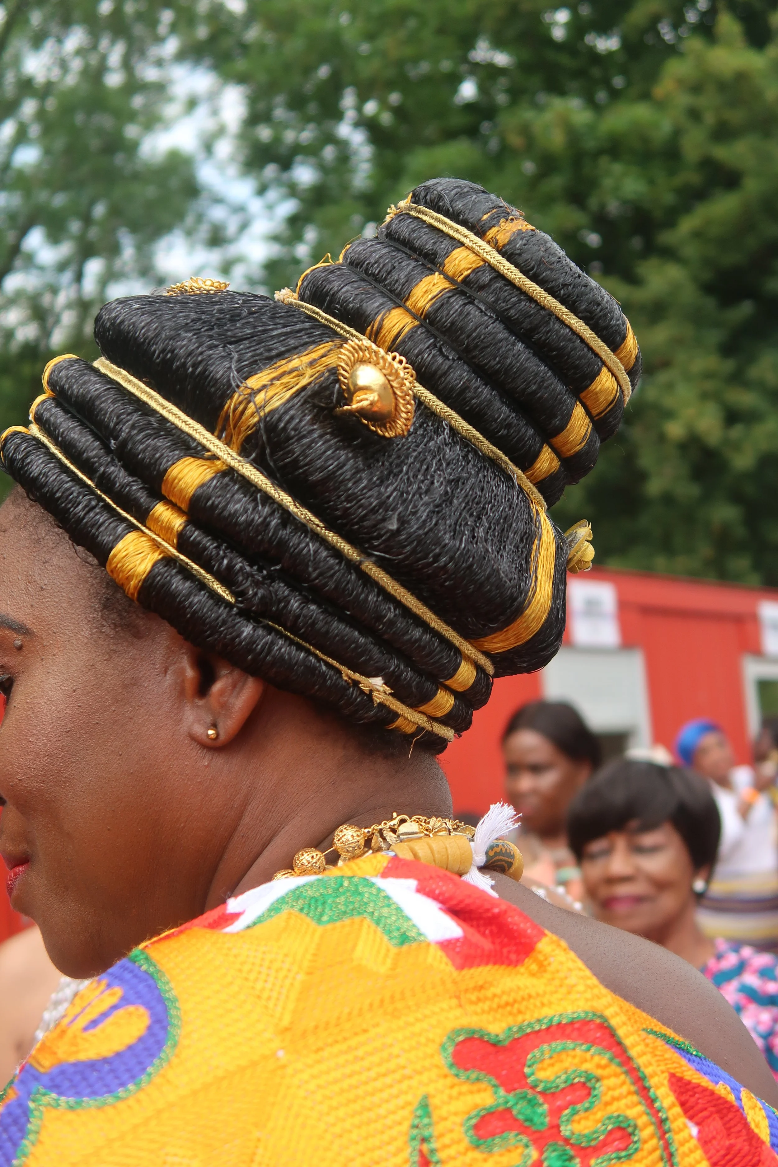 A woman wearing a black and gold traditional headdress with gold jewelry, seen from the side, outdoors with people in the background.