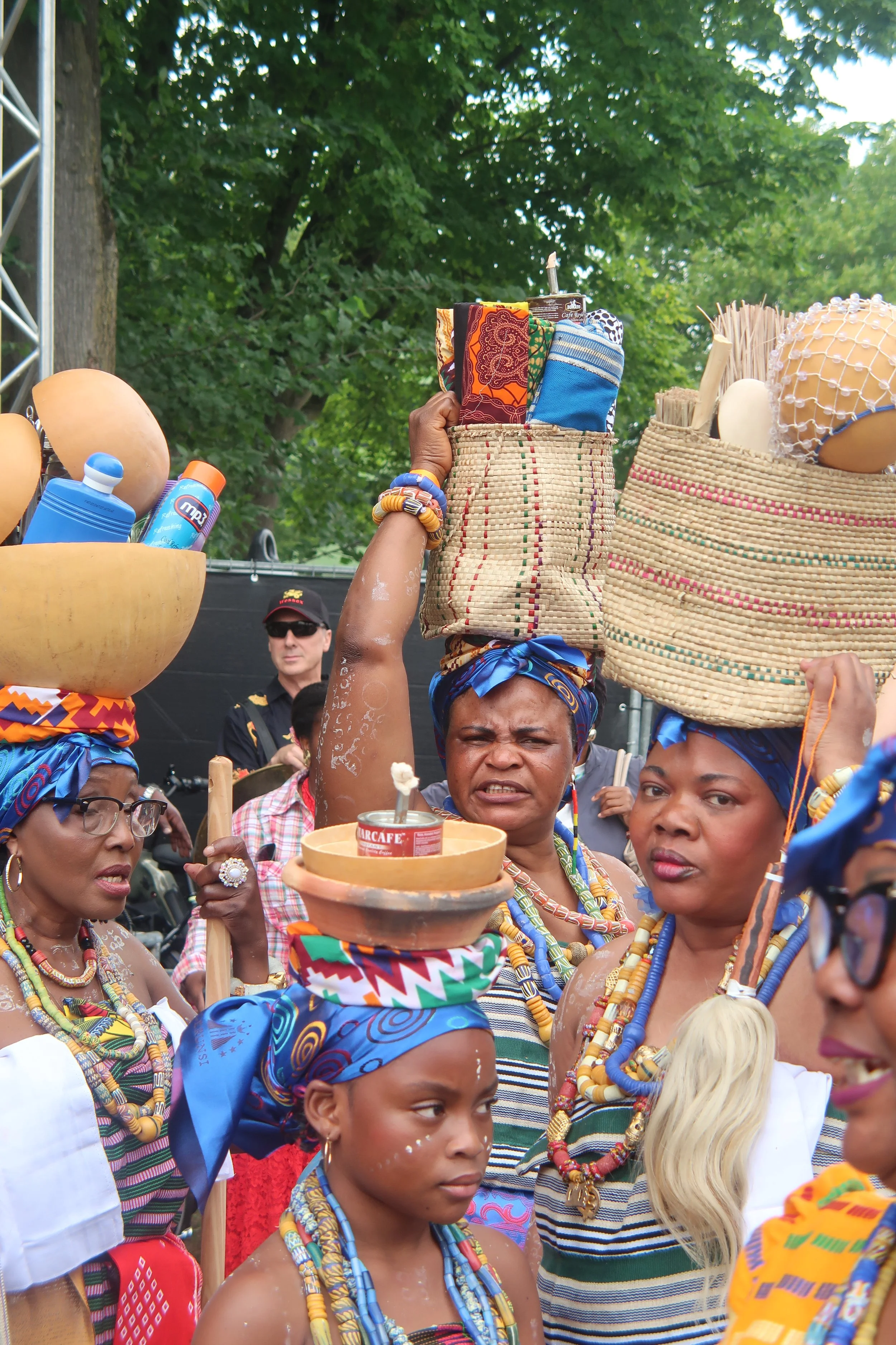 A group of women dressed in traditional African attire and jewelry participating in a cultural event or celebration outdoors on a sunny day, with some carrying baskets on their heads and one woman holding a tray with a can of tomato sauce.
