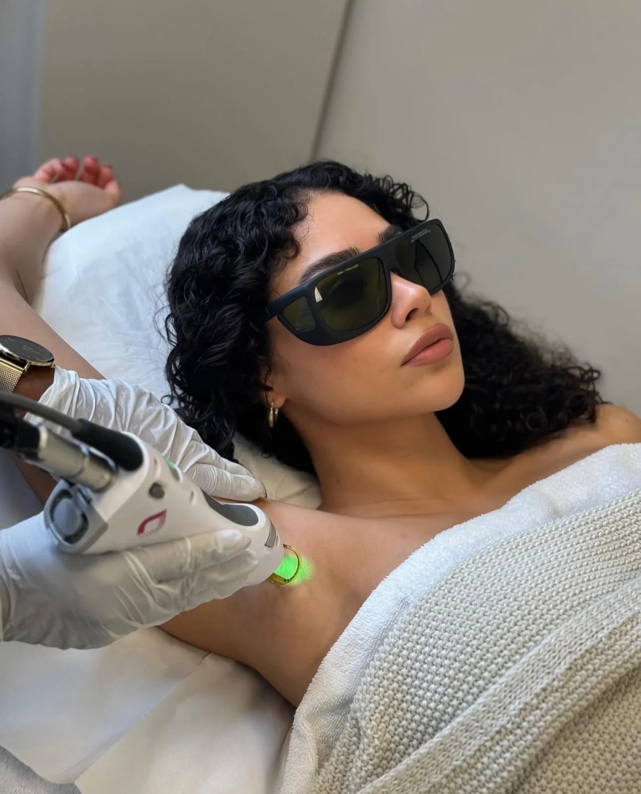 Woman with curly hair wearing sunglasses receiving laser treatment on her underarm in a medical facility.