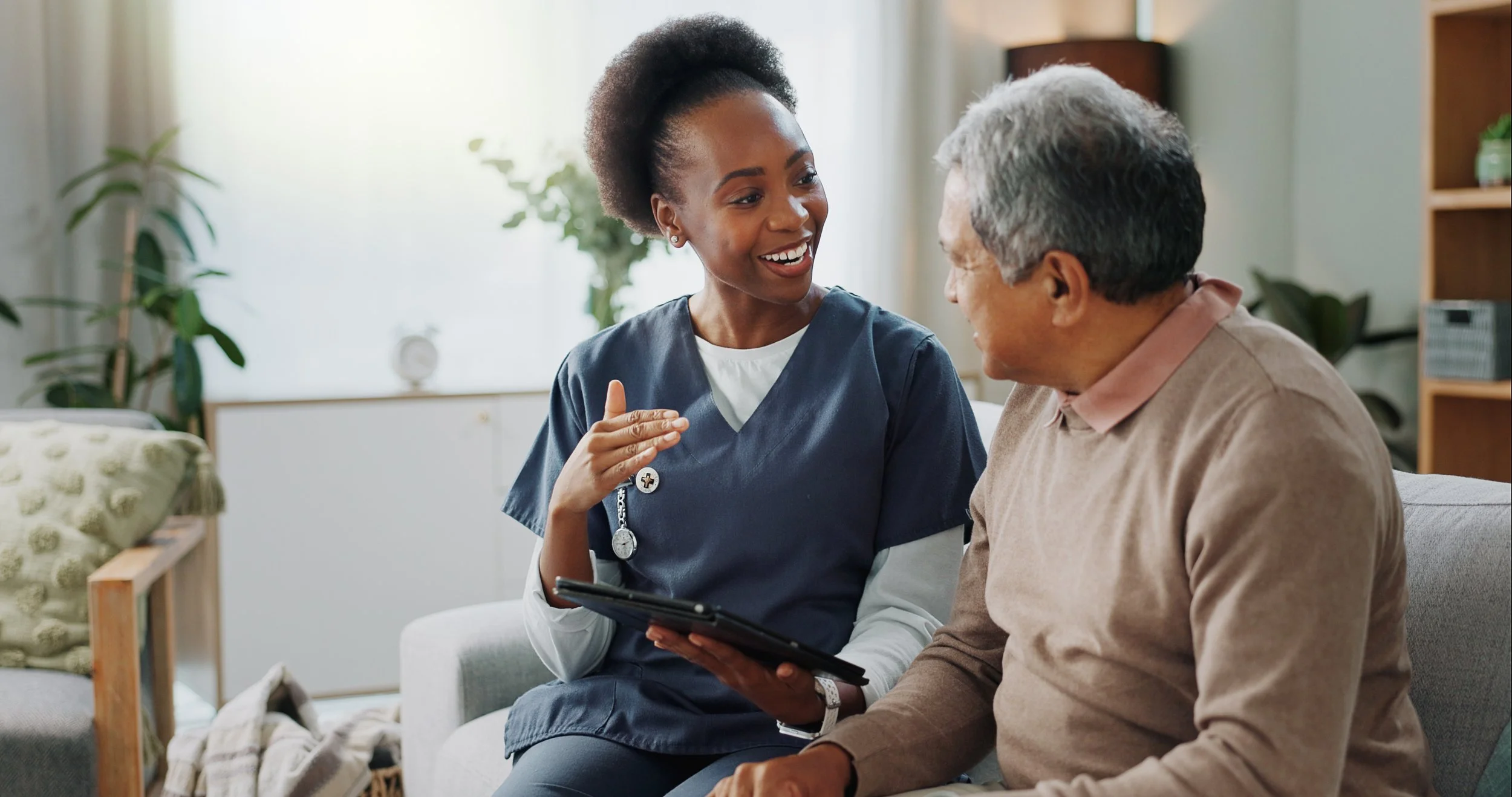 A young female nurse in blue scrubs talking to an elderly man in a cozy living room.