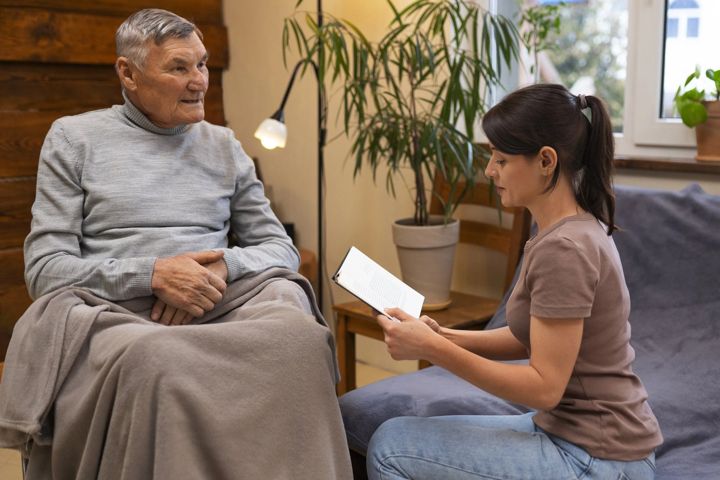 A young woman is sitting on a chair, reading a document, and talking to an elderly man sitting across from her. They are in a cozy room with a large window, a potted plant, and warm lighting.