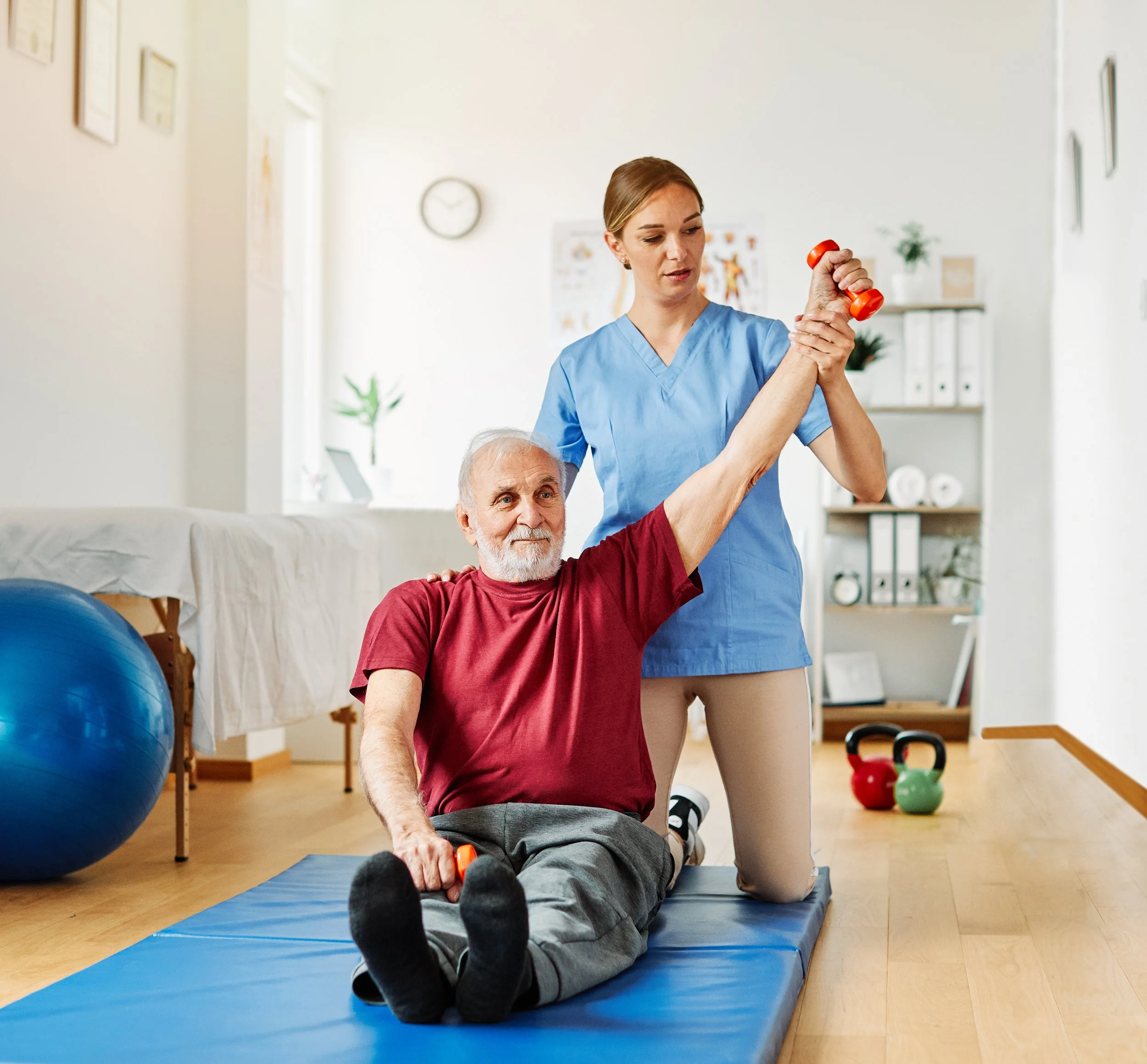 A healthcare professional in blue scrubs assisting an elderly man with arm exercises using small dumbbells during physical therapy in a recovery room.