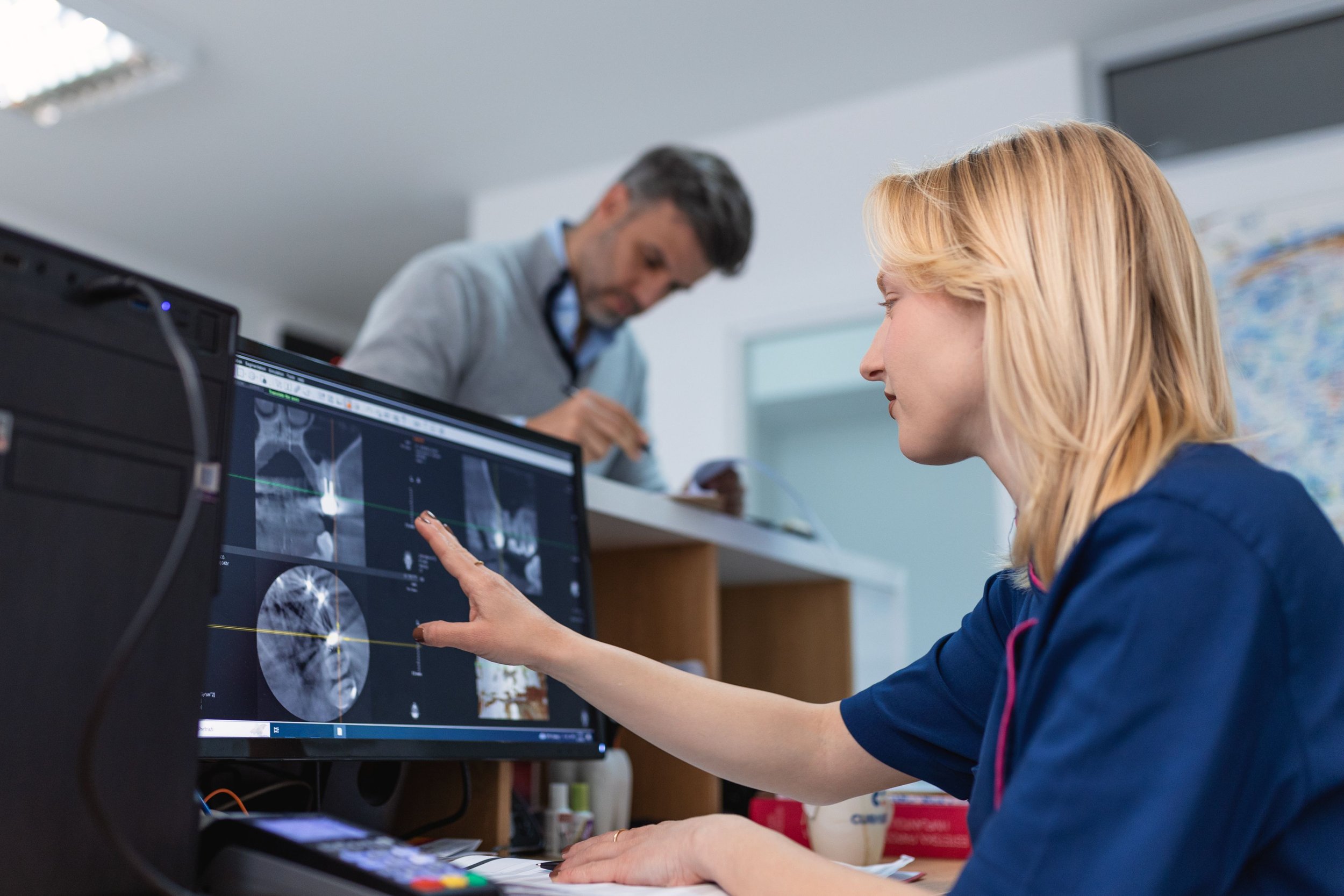 A woman in medical scrubs analyzing brain scans on a computer monitor in a healthcare setting, with a man in the background writing at a counter.