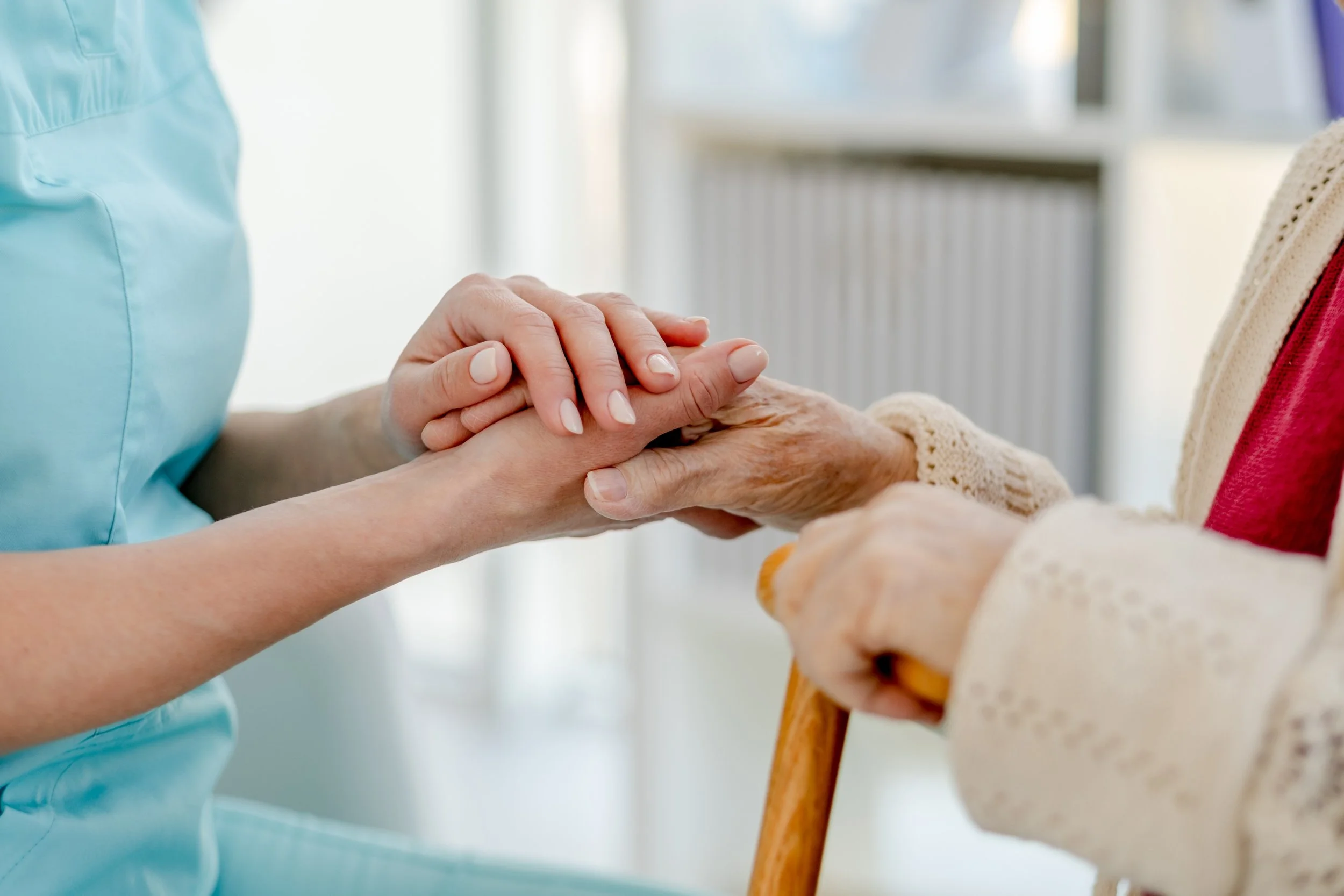 A caregiver holding an elderly woman's hand, who is sitting with a walker, in a caring gesture.