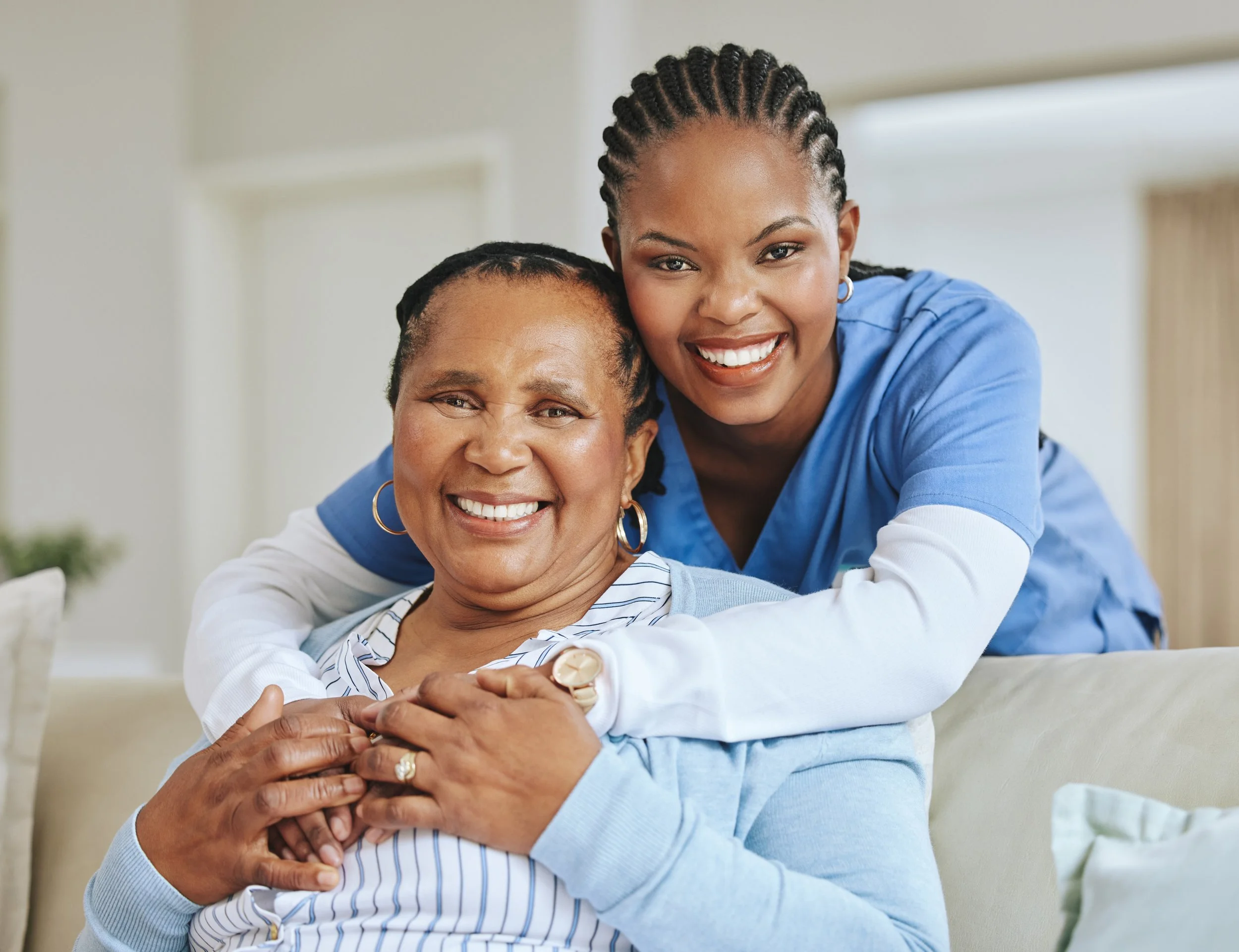 A young woman with braids hugs an older woman with short hair and hoop earrings, both smiling warmly in a cozy, well-lit room.