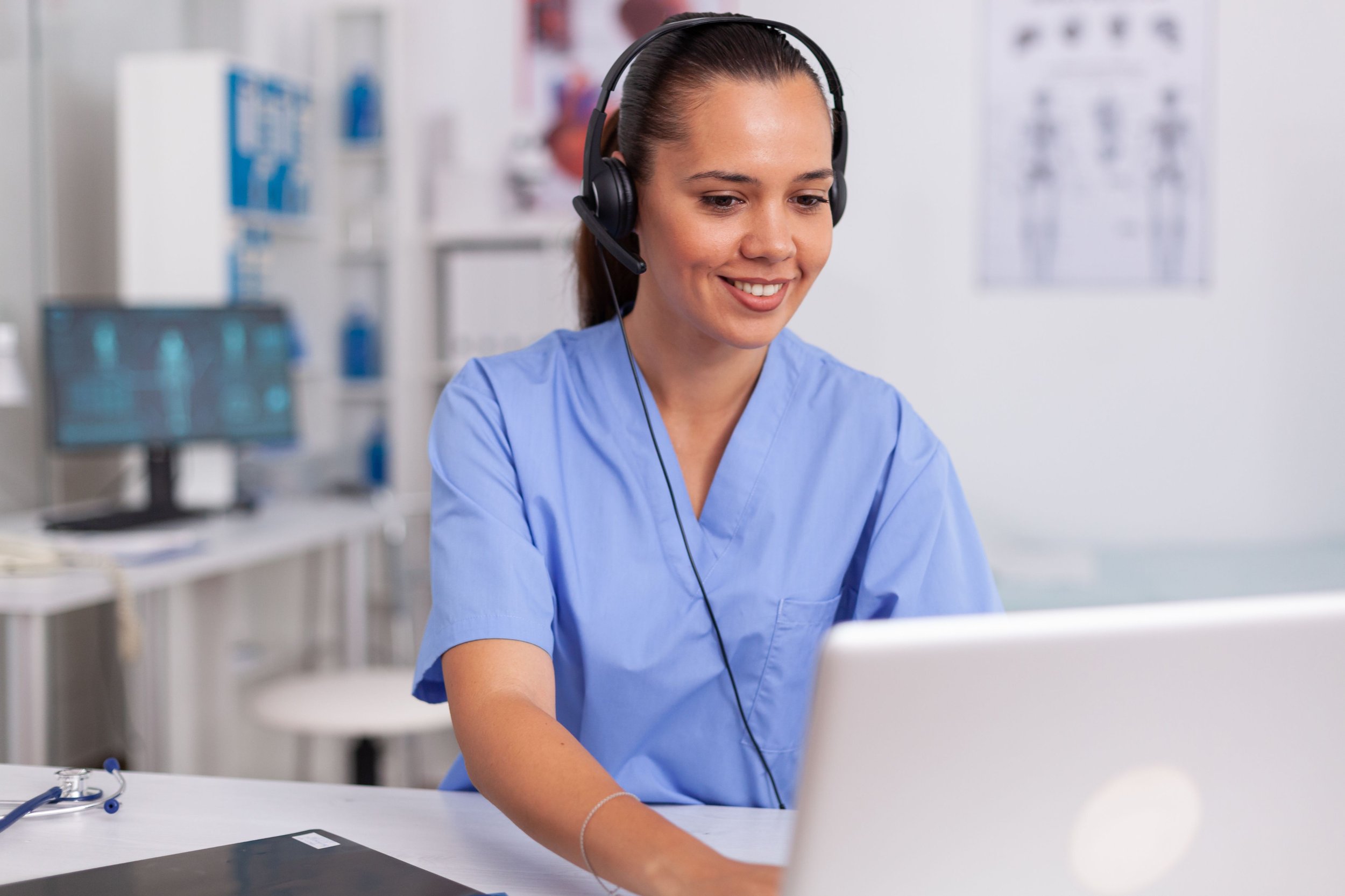 A young woman wearing blue scrubs and a headset sitting at a desk in a medical or office setting, smiling and working on a computer.