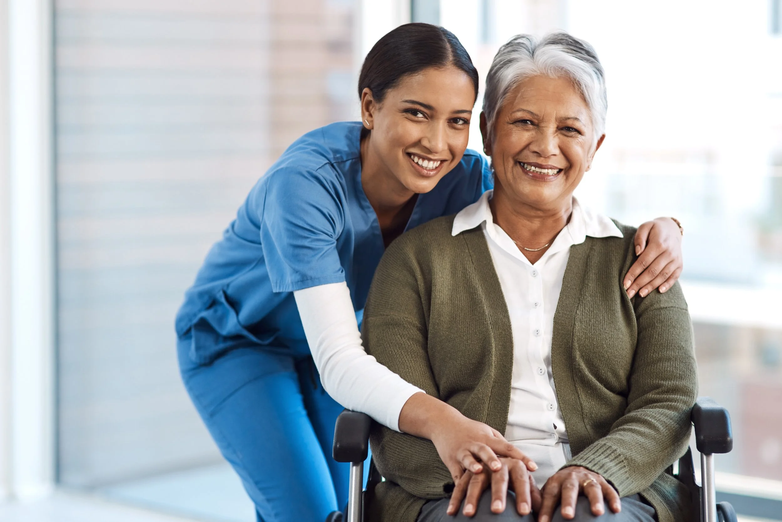 A healthcare worker in blue scrubs smiling and hugging an elderly woman with white hair, who is sitting in a wheelchair, in a bright room.