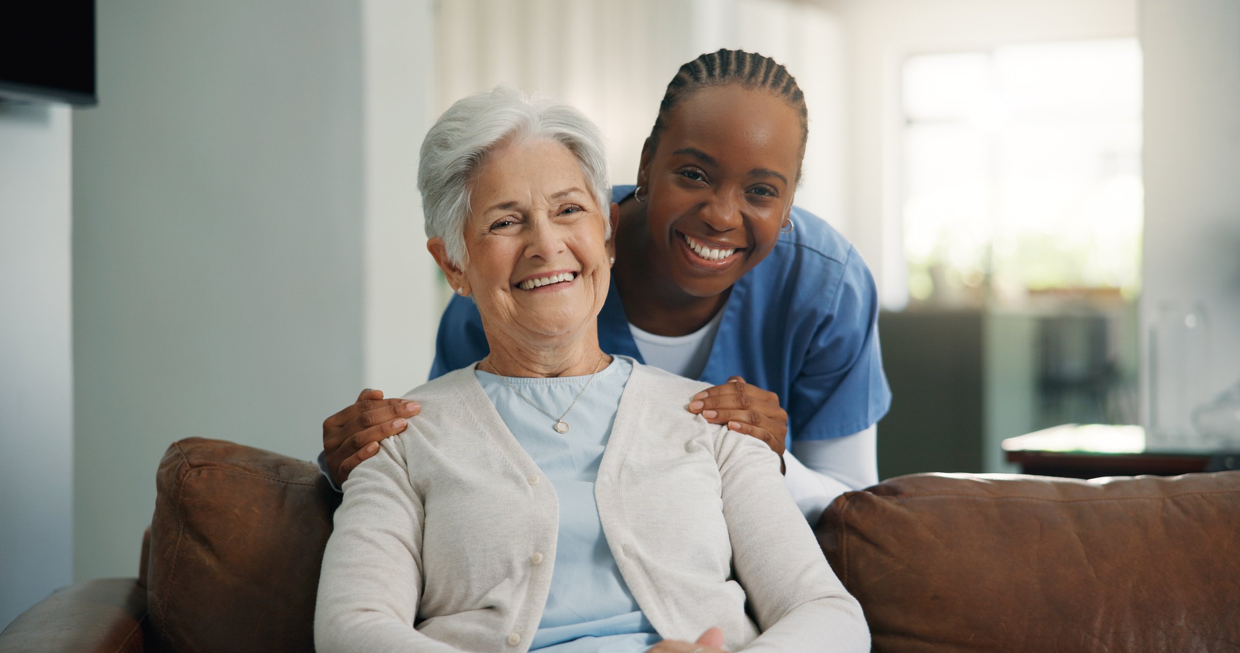 A young African American caregiver in blue scrubs smiling and hugging an elderly Caucasian woman with gray hair sitting on a brown couch in a bright, modern living room.