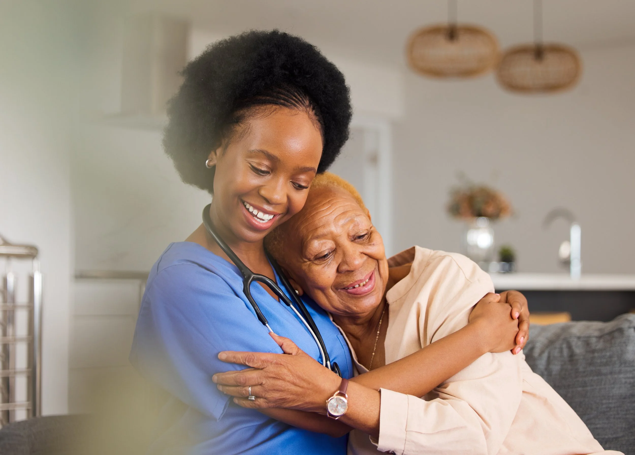 A healthcare professional, wearing blue scrubs and a stethoscope, hugs an elderly woman with grey hair, who appears to be content and happy in a home or medical setting.