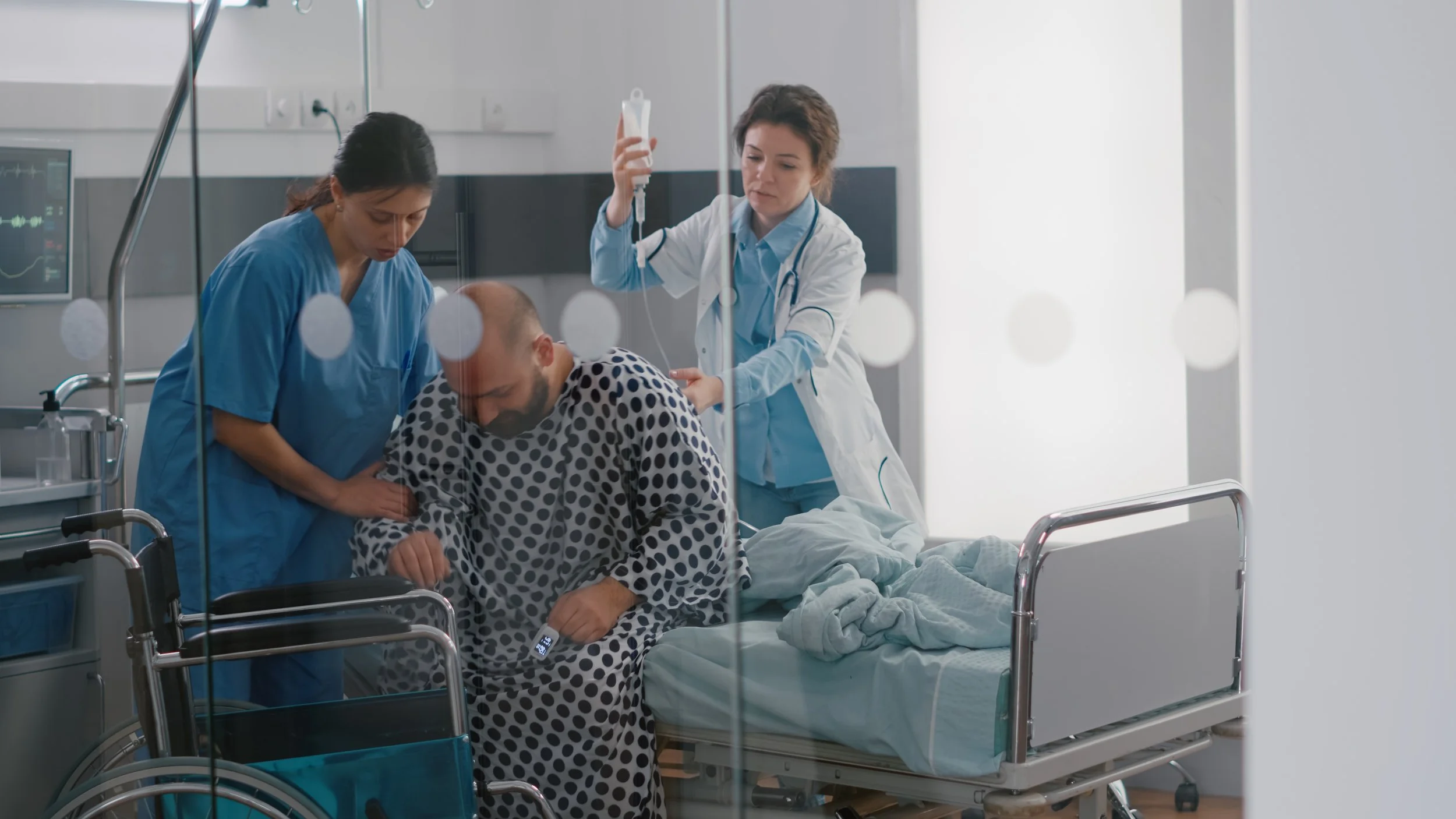 Healthcare professionals attending to a patient in a hospital room. The patient appears to be a man in a hospital gown, sitting on a bed with medical equipment nearby. One healthcare worker in blue scrubs is leaning over him, while another in a white coat is hanging an IV bag.