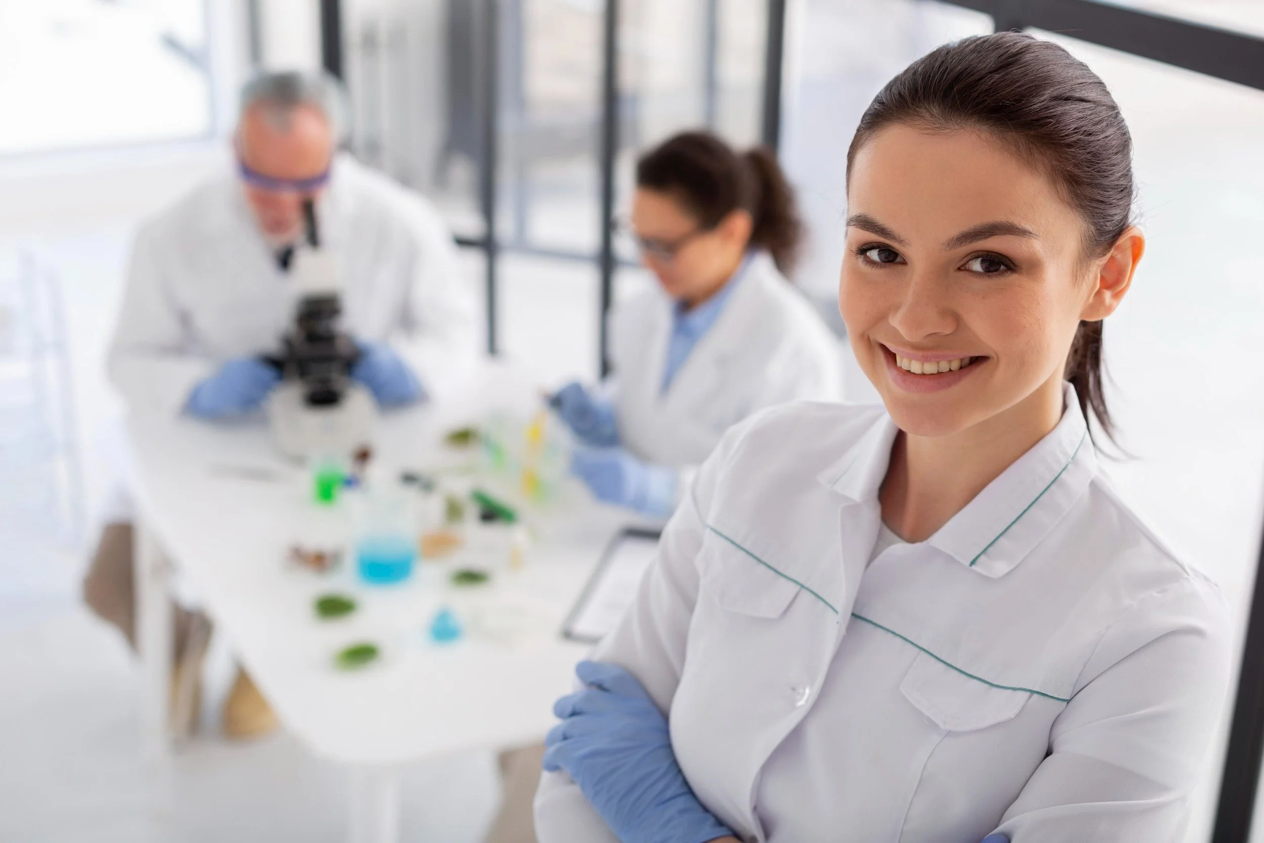 Smiling female scientist in lab coat and gloves, with two colleagues working with a microscope and lab equipment in the background.