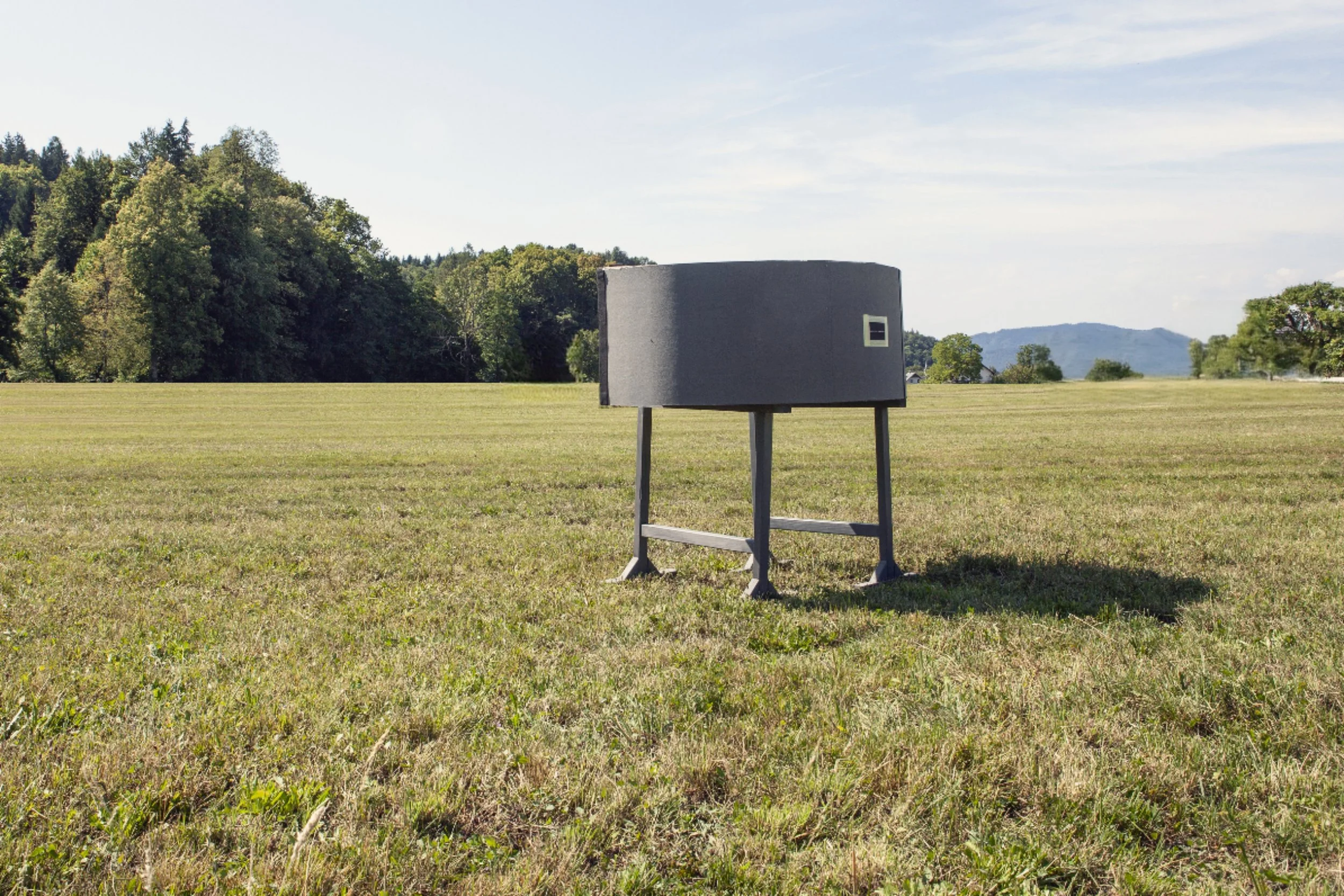 A portable camera obscura in the shape of a half-cylinder,  in an open grassy field with trees and hills in the background.