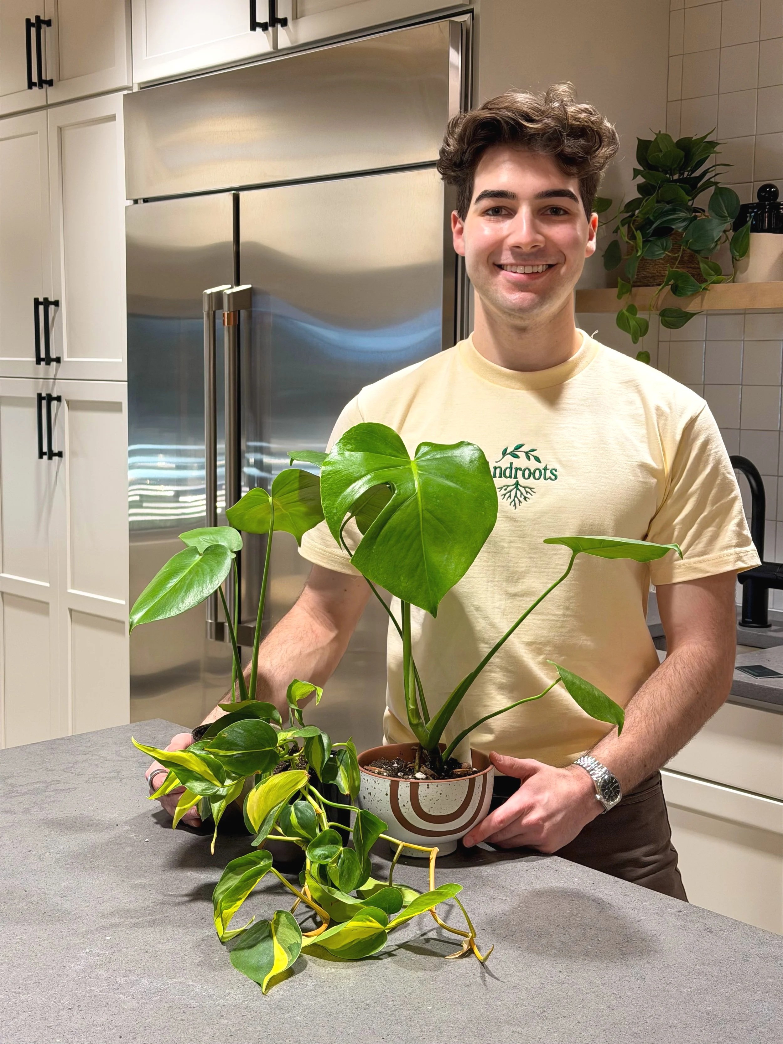 A young man with curly brown hair smiling while holding a potted houseplant with large green leaves, standing in a modern kitchen with white cabinets and stainless steel appliances.