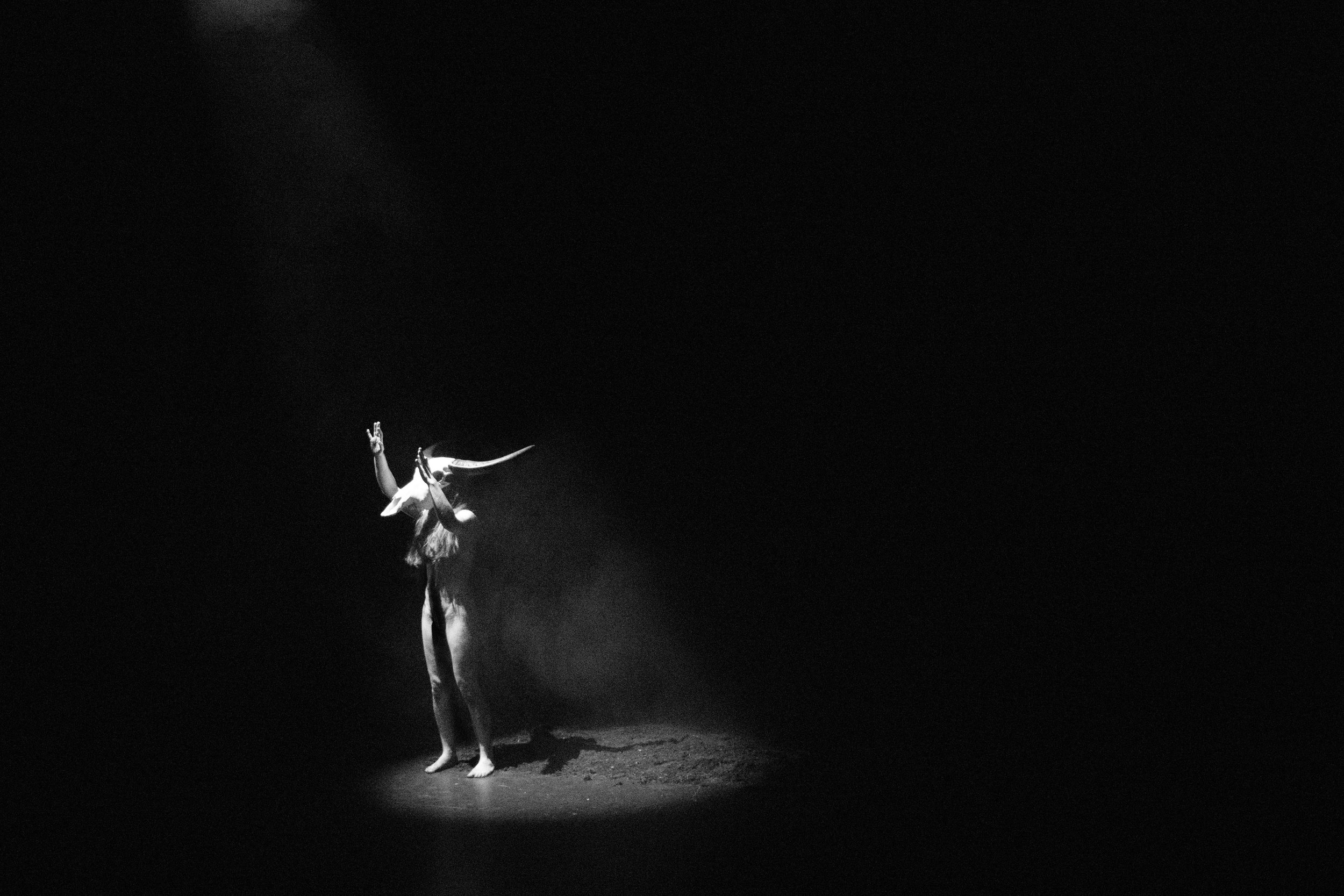 A performer stands alone on stage illuminated by a spotlight in a dark setting, wearing a costume with an elaborate headpiece or horns and making a peace sign gesture with their right hand.