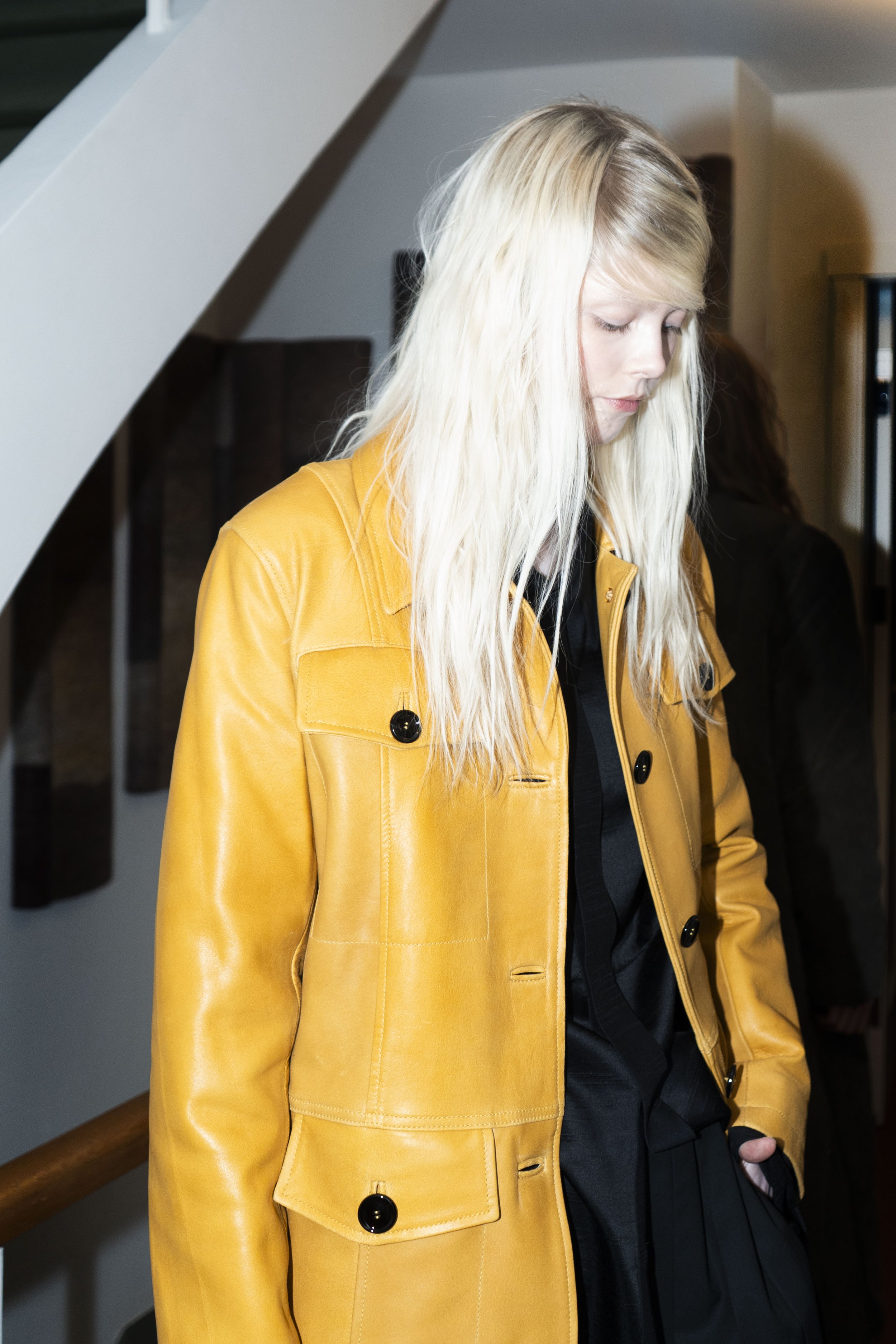 A woman with long blond hair wearing a yellow leather coat, looking down with her hands in her coat pockets, indoors near a staircase.