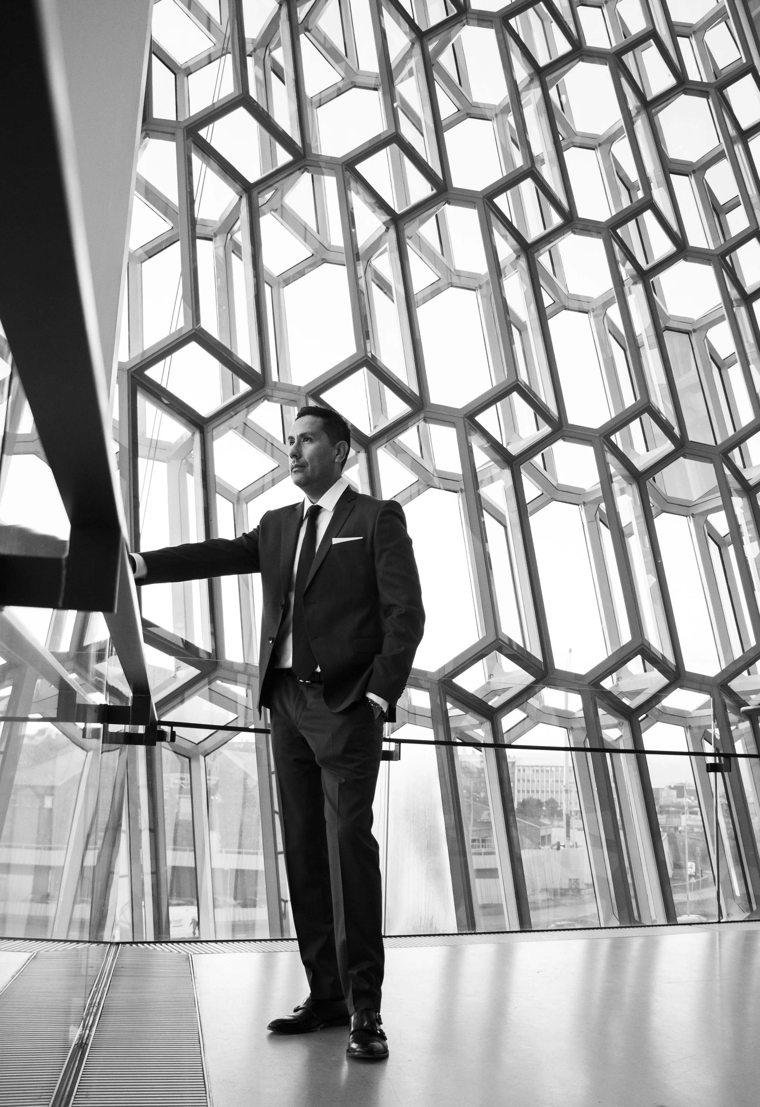 A man in a black suit standing inside a modern building with a large geometric glass facade, looking thoughtfully outside.